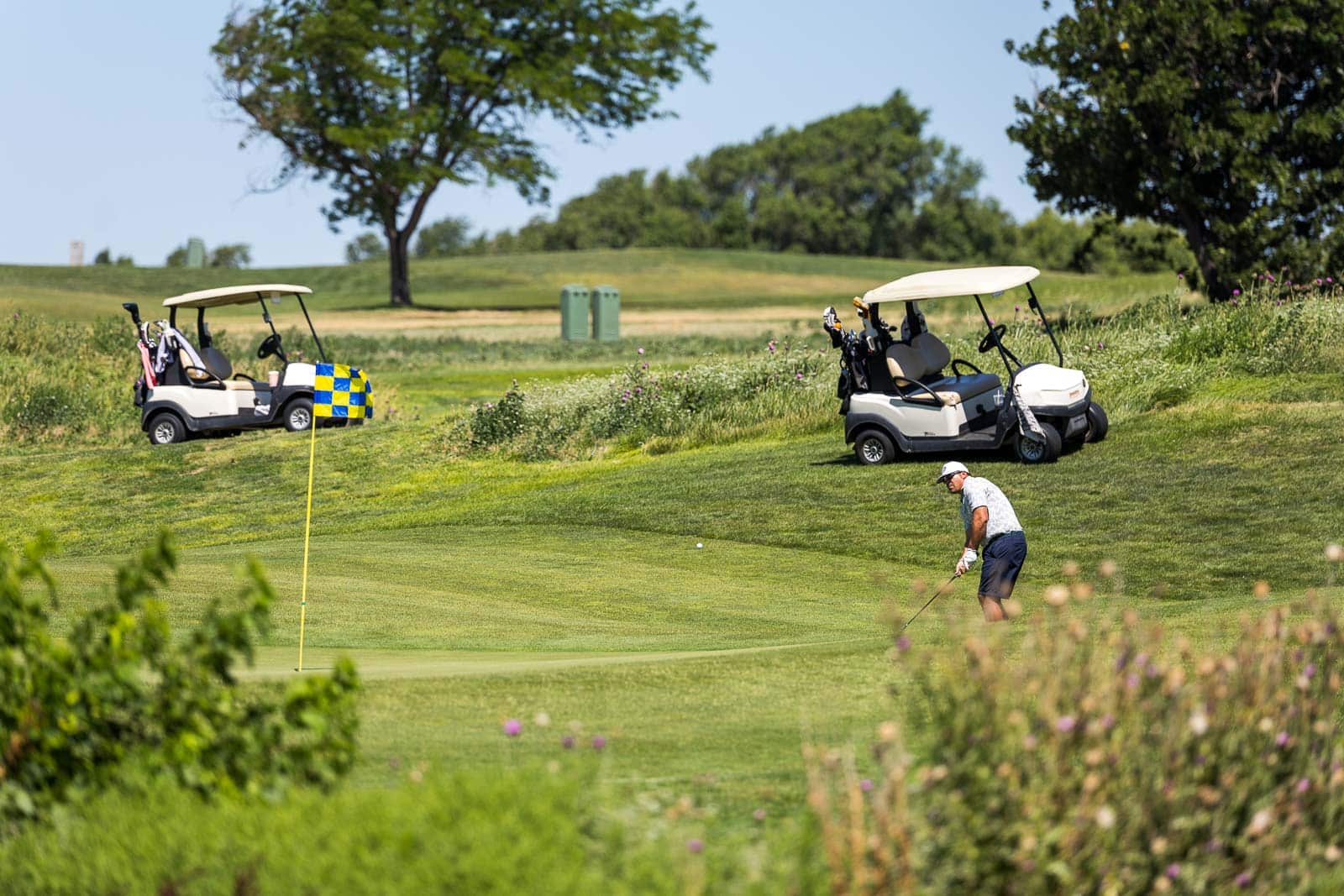 Golfer in a white shirt and navy shorts taking a swing on a sunny golf course with two golf carts nearby on the grassy hilltop in the background, flagpole in foreground.