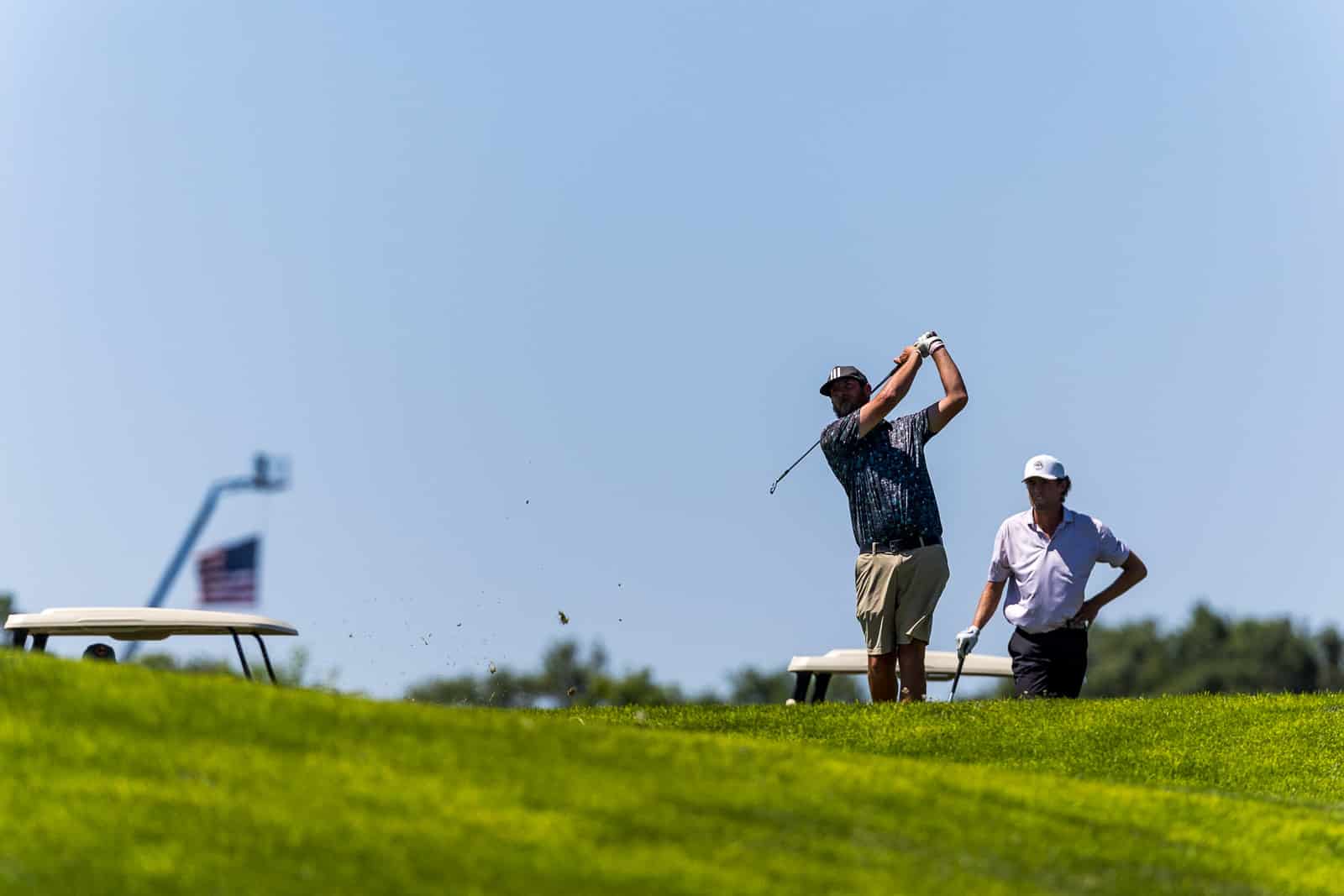 Two men on a sunny golf course; one man is swinging a club while the other watches, with a golf cart in the distance and a clear blue sky above.