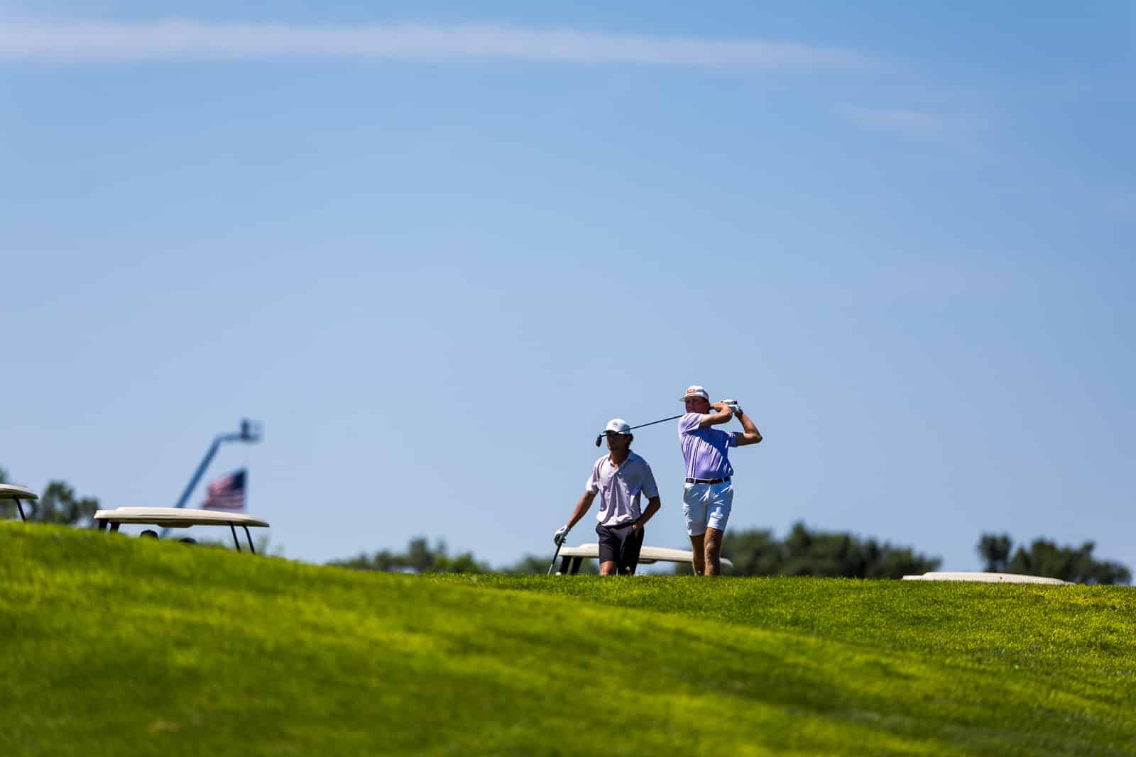 Two men playing golf on a sunny course: one golfer is mid-swing while the other stands nearby watching on a grassy hill under a clear blue sky.