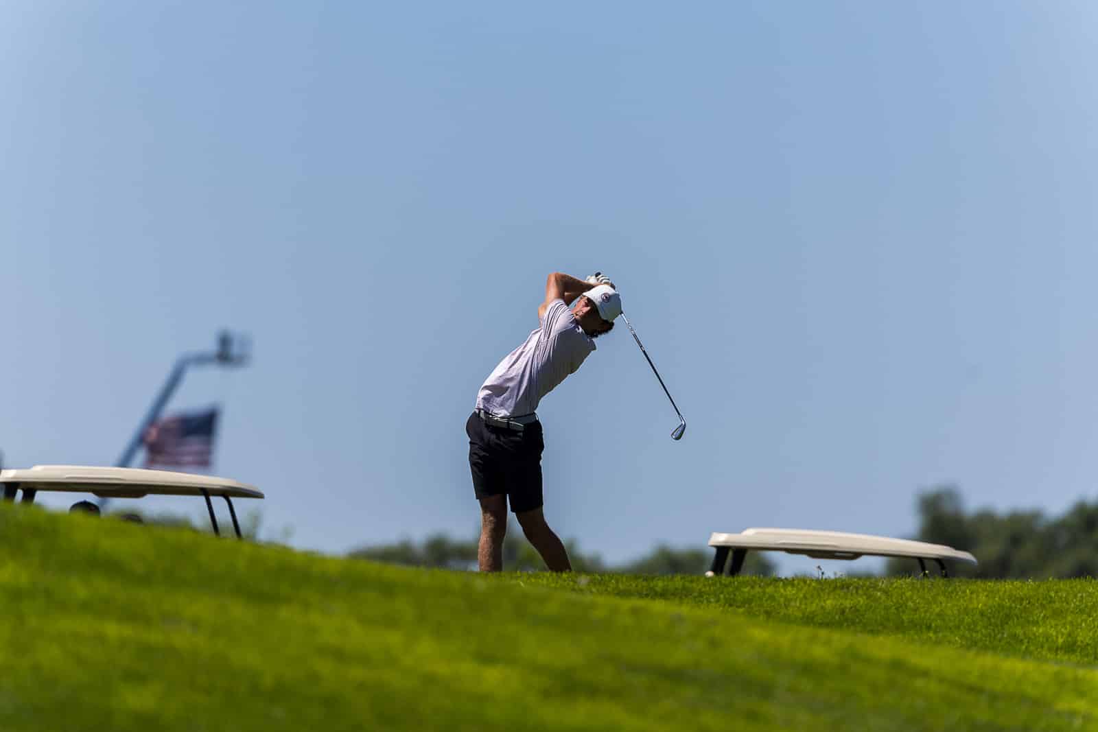 Golfer in a backswing on a sunny golf course, blue sky overhead and carts in the distance.