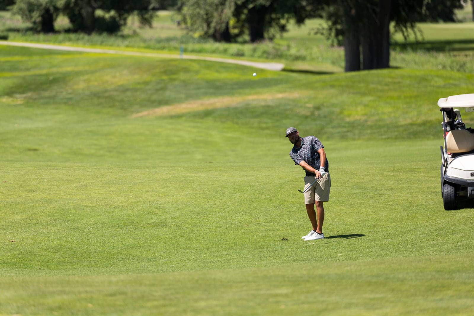 Golfer in a patterned shirt mid-swing on a sunny, grassy golf course with a golf cart parked to the right.