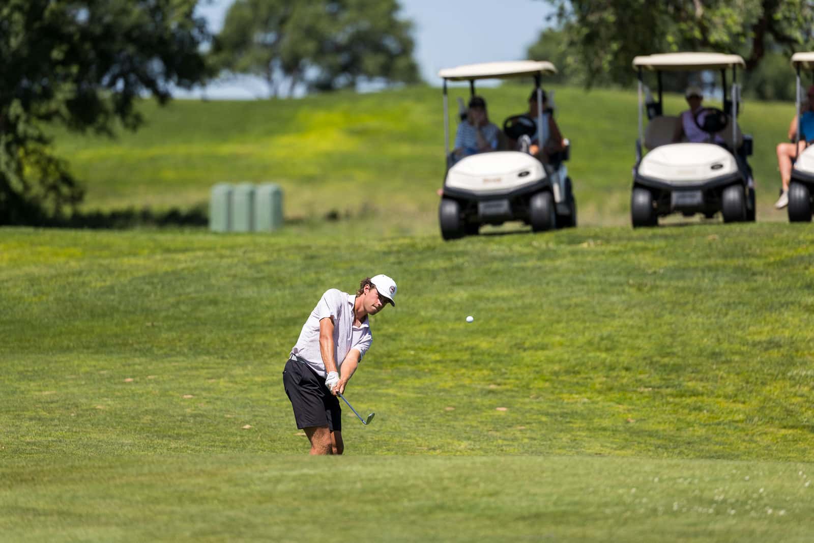 Golfer in a white shirt and cap swings on a lush golf course with golf carts in the background.