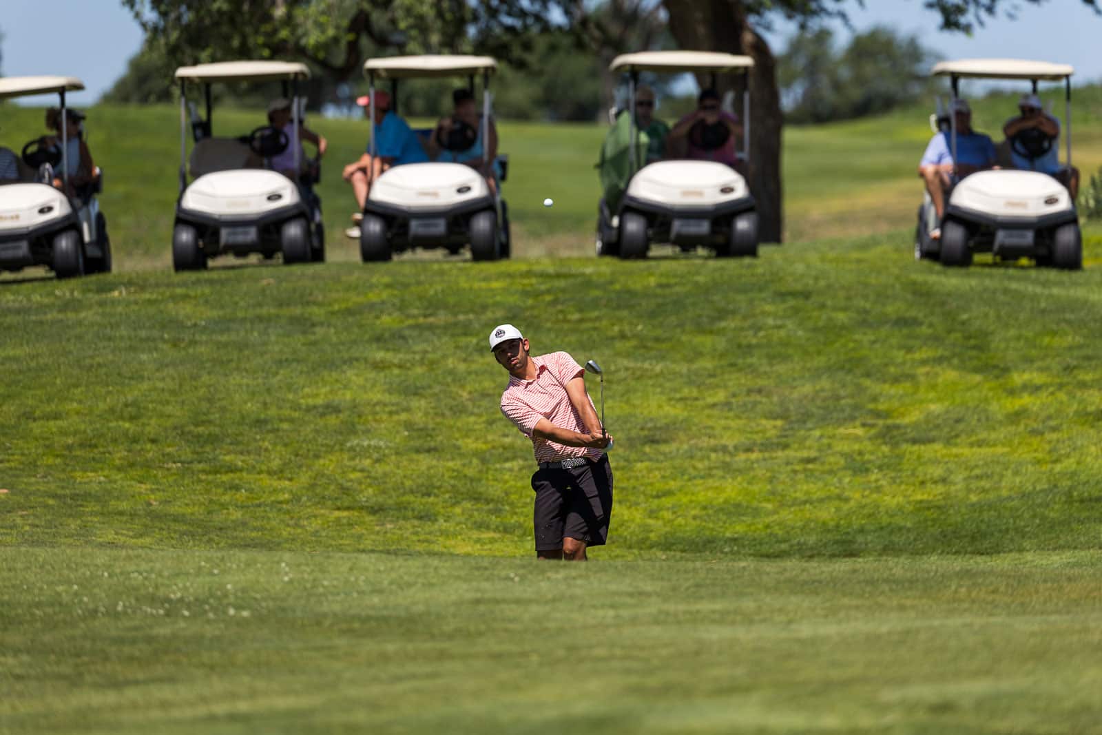 Golfer in a pink striped shirt and white cap mid-swing on a sunny golf course, with spectators in golf carts behind him.