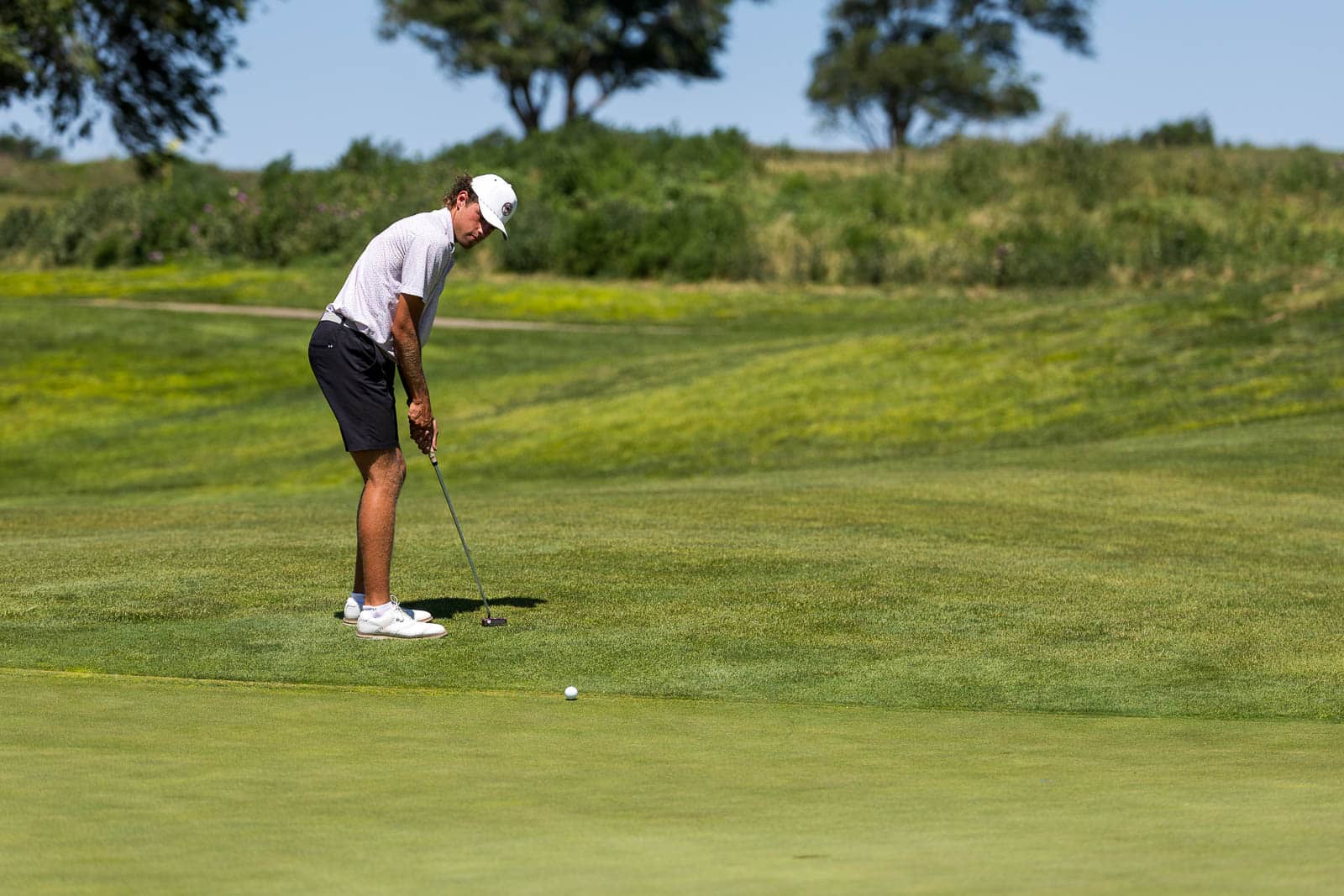Male golfer in a white cap and light shirt prepares a short putt on a bright green golf course green.