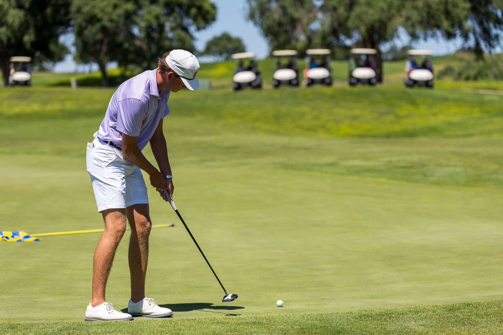 Male golfer in a lavender polo and white shorts prepares to hit a shot from the green, with a ball near the putter on a sunny course and golf carts in the distance.