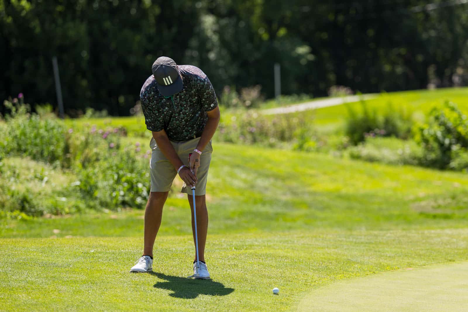 Golfer preparing to putt on a green, wearing a dark cap and floral shirt, focused on the ball nearby.