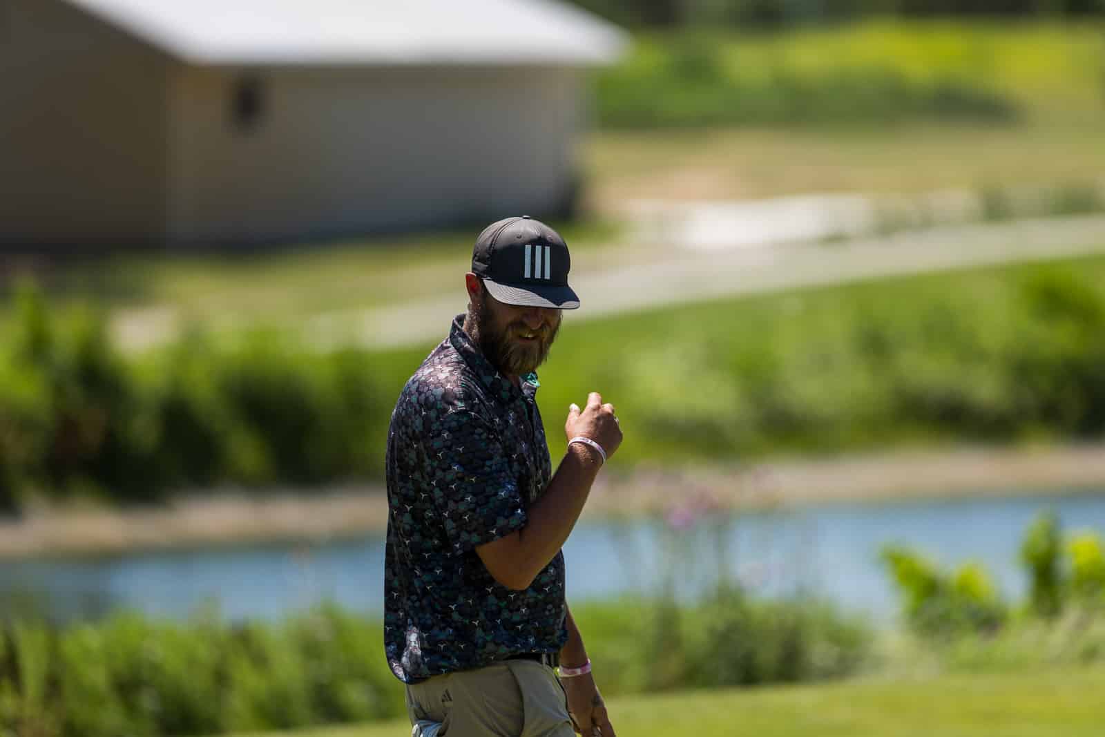 Bearded man in a black cap and patterned shirt stands outdoors near a lake.