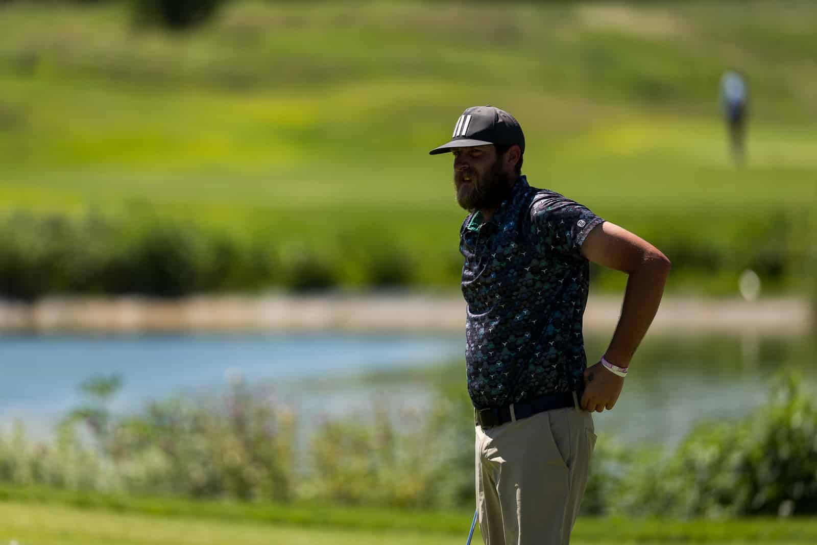 Bearded man in a dark cap and patterned polo stands with hands on hips by a lake.