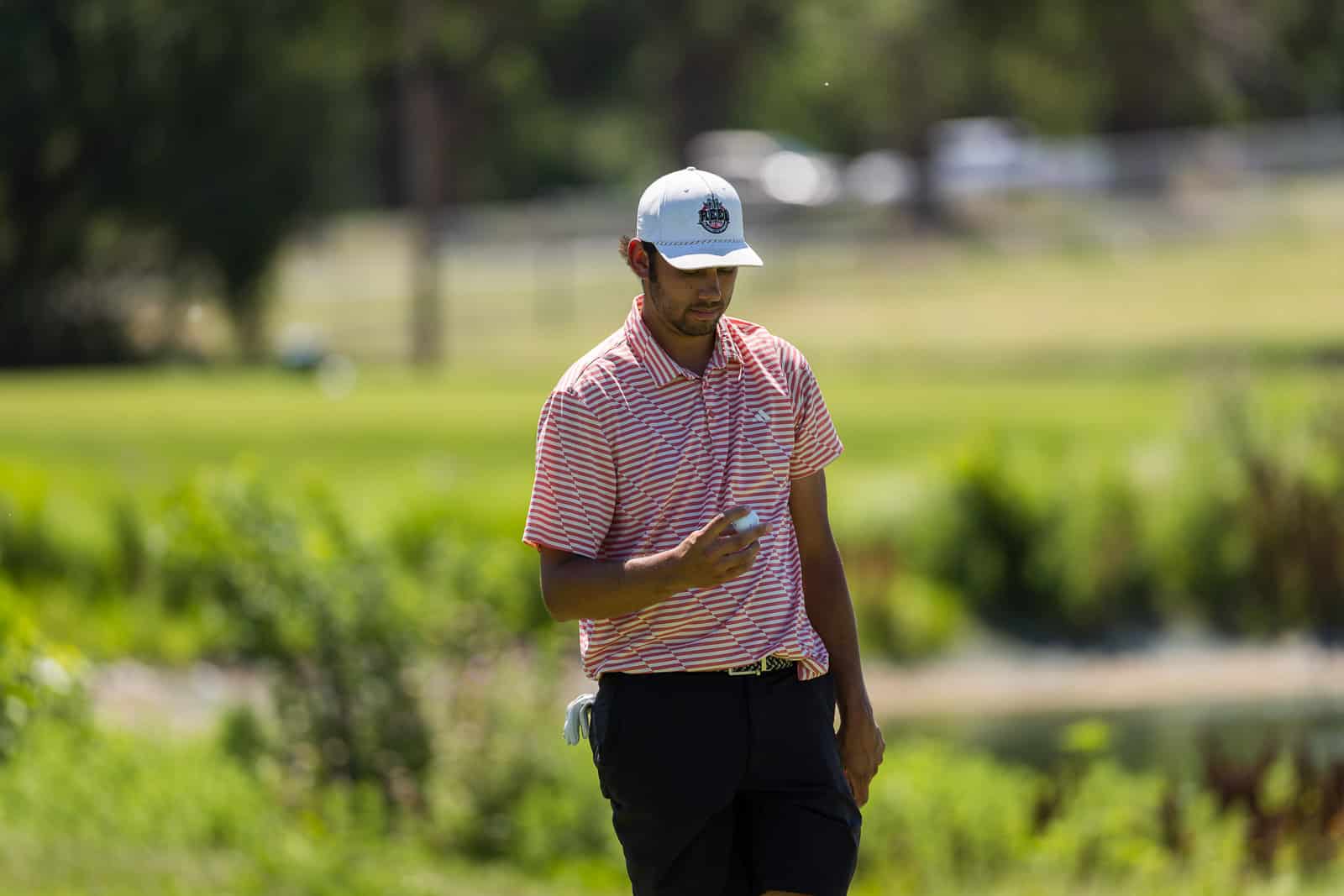 A man in a red and white striped polo shirt holding a golf ball on a sunny golf course.
