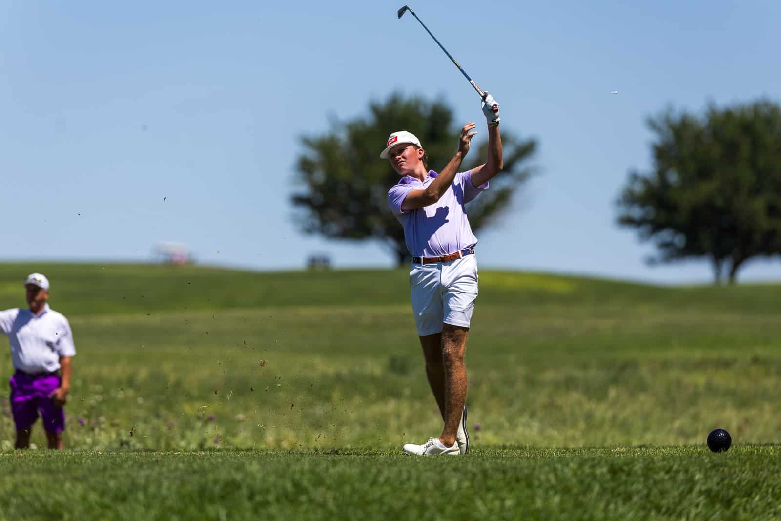 Golfer in lavender polo and white shorts mid-swing on a sunny golf course, with a spectator in white and purple in the background.