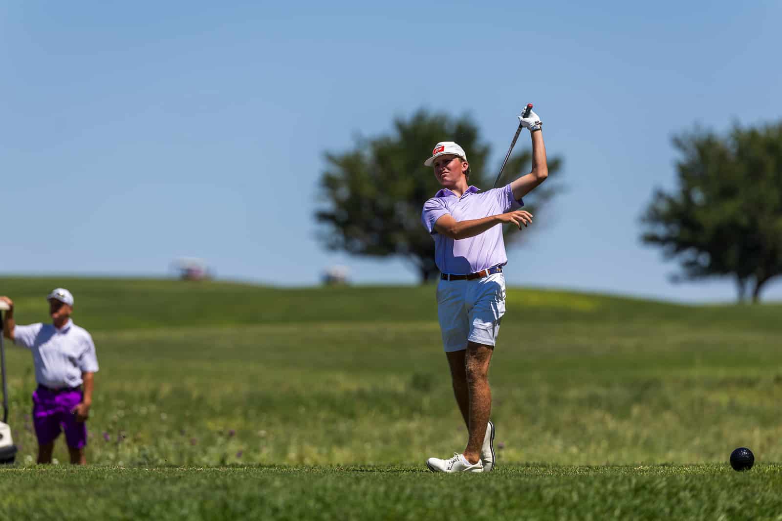 Golfer in lavender polo and white cap mid-swing on a sunny golf course, with a spectator in the background.