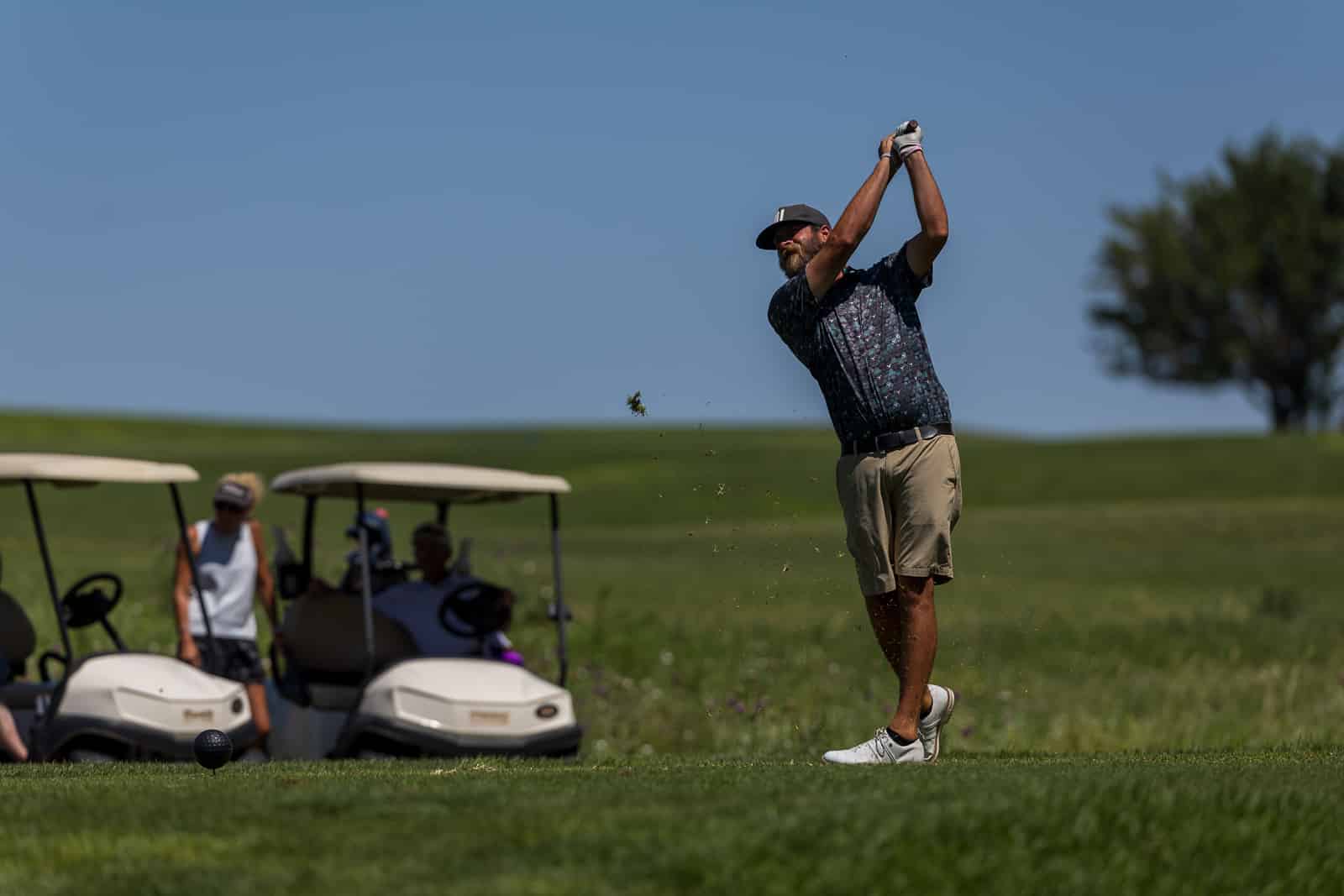 Golfer in a patterned shirt and beige shorts mid-swing on a sunny golf course, with spectators and golf carts in the background.