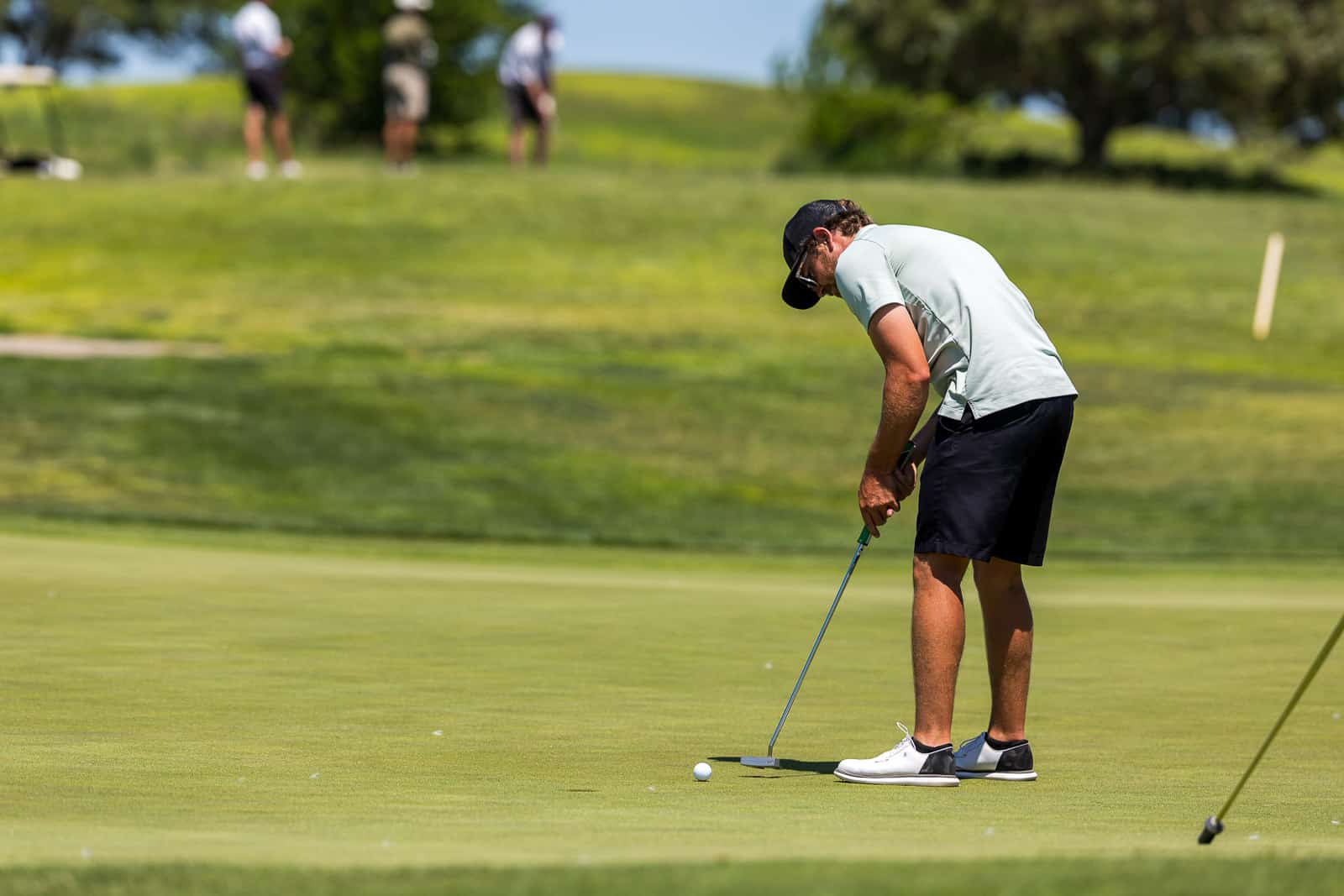 Golfer preparing to putt on a manicured green, focusing on the ball with a putter in hand under sunny skies.