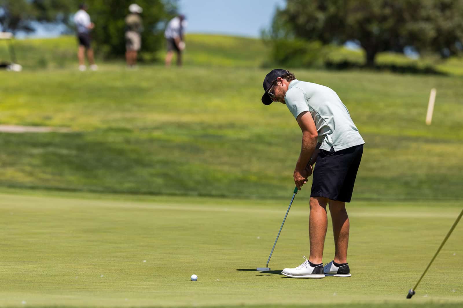 Golfer in light gray shirt and black cap lining up a putt on a green, with players on a hill in the blurred background.