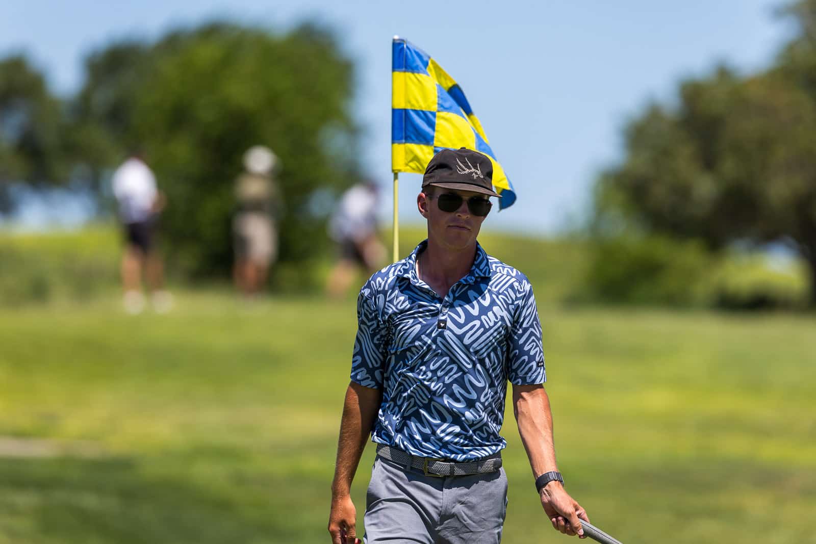 Male golfer standing on a sunny golf course near a yellow-and-blue flag, wearing a patterned blue shirt and dark sunglasses.
