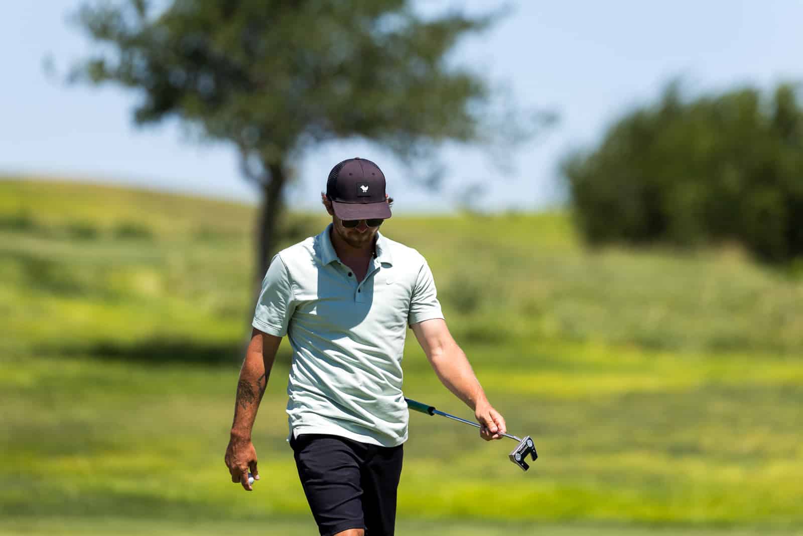 Man in a light polo and cap walks on a sunny golf course, holding a golf club and wearing sunglasses.