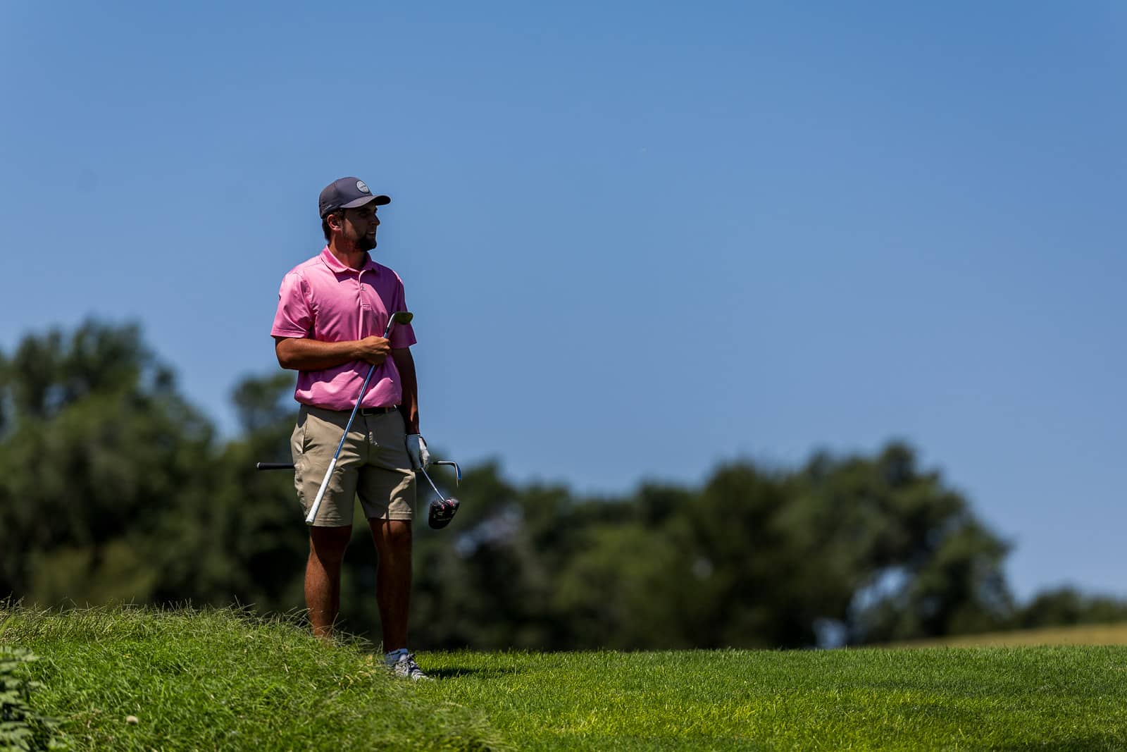 Golfer in a pink polo and cap stands on a sunny golf course, holding a club and looking into the distance.