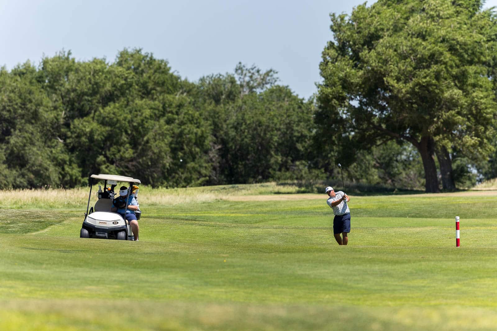 Golfer mid-swing on a sunny golf course while a golf cart with two players sits nearby on the fairway.