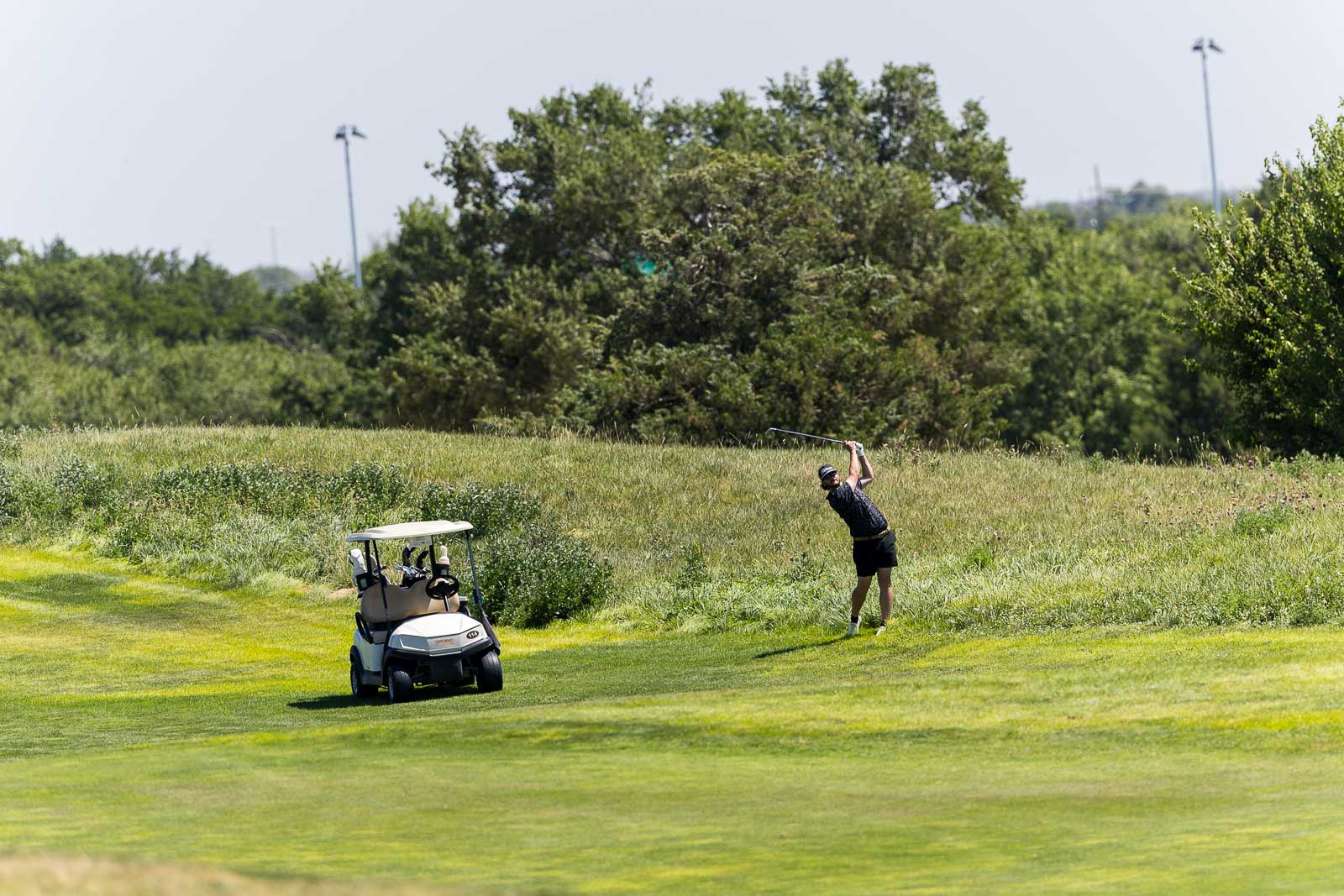 Golfer mid-swing on a lush golf fairway with a golf cart nearby.