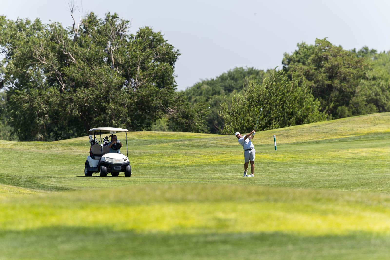Golfer in light clothes hits ball on a sunny green golf course while a golf cart with bags sits nearby.