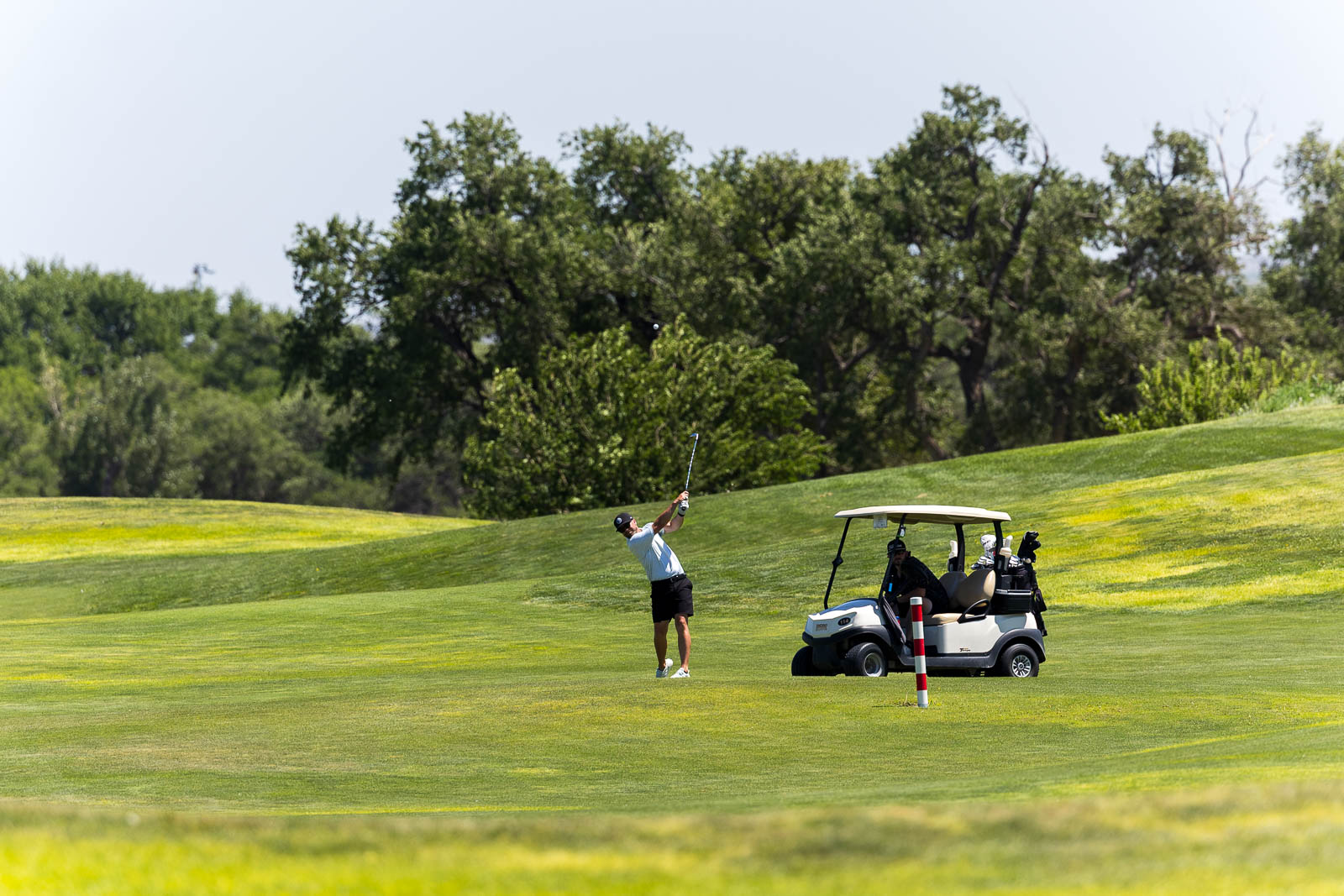 Golfer mid-swing on a sunny golf course with a parked golf cart and clubs nearby on the fairway