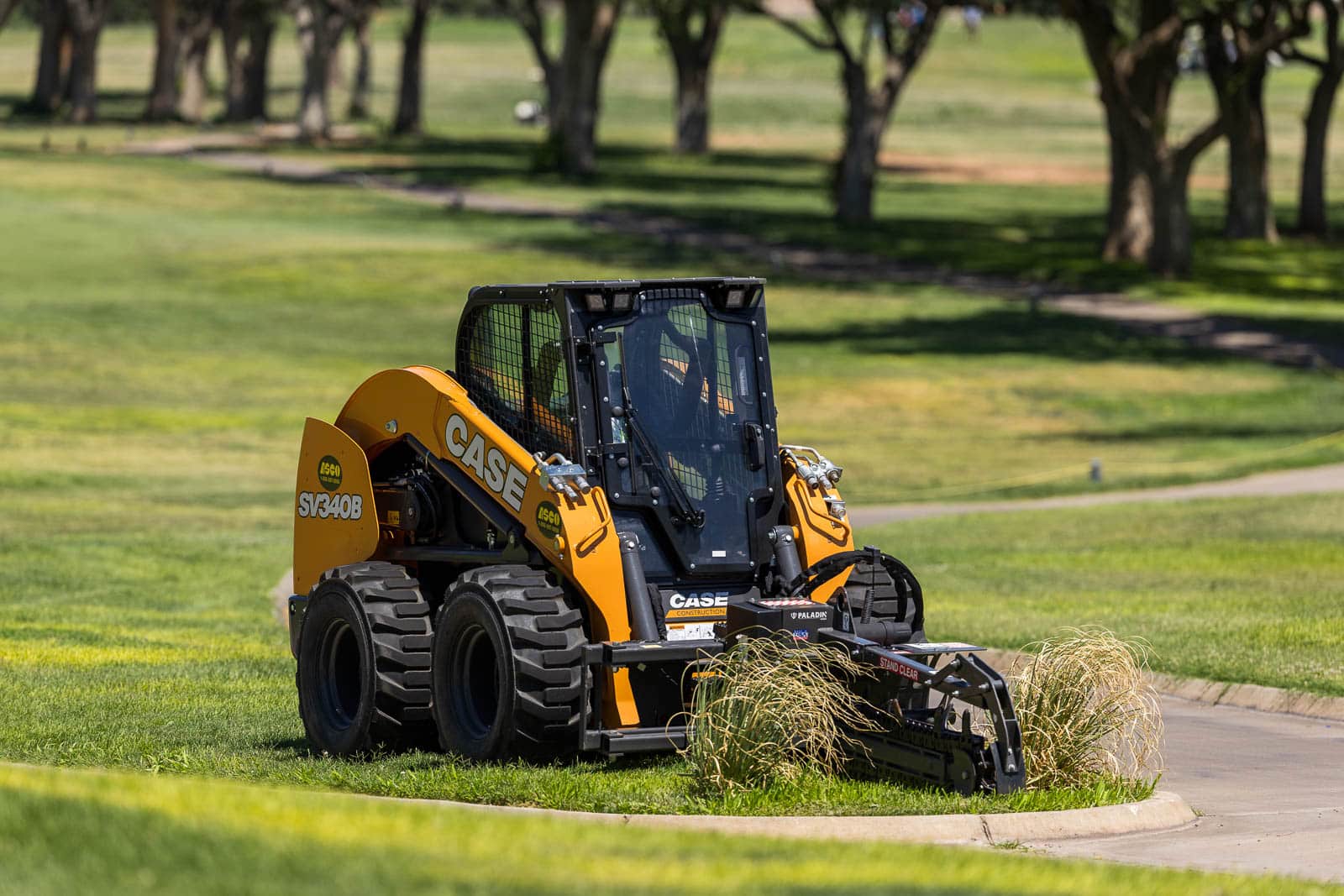 Orange CASE skid-steer loader with a green landscape rake attachment parked on a grassy park area, with trees in the background