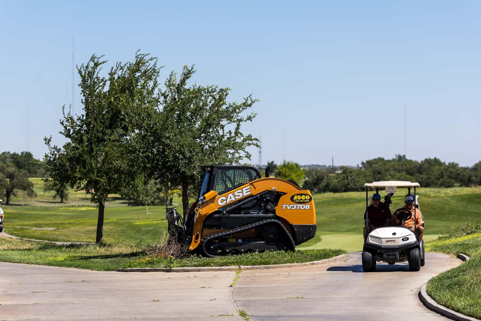 Orange CASE compact excavator parked on a golf course path beside a tree, with a two-person golf cart in the background