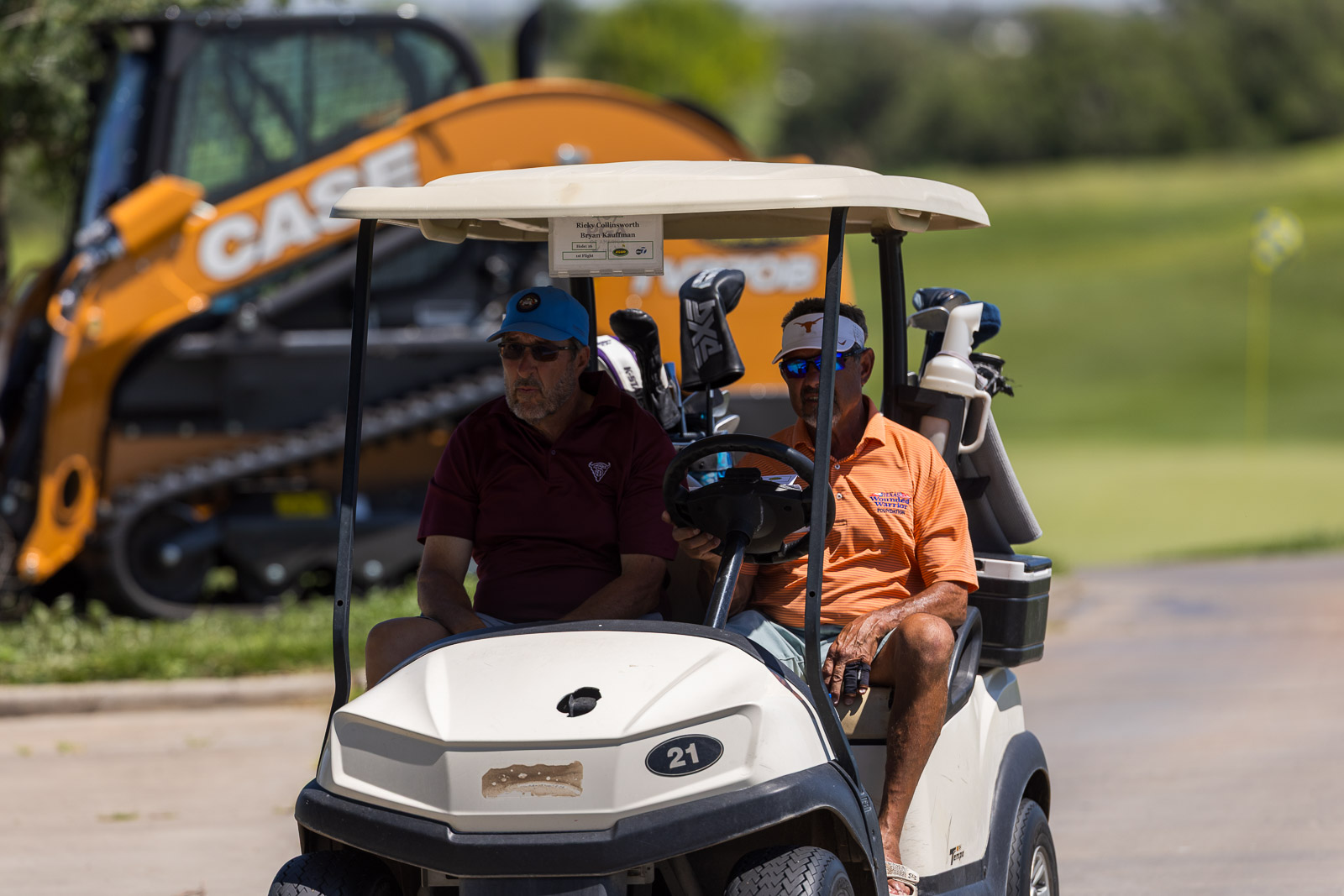 Two men ride in a golf cart on a sunny course with golf bags behind them and a large orange excavator blurred in the background.