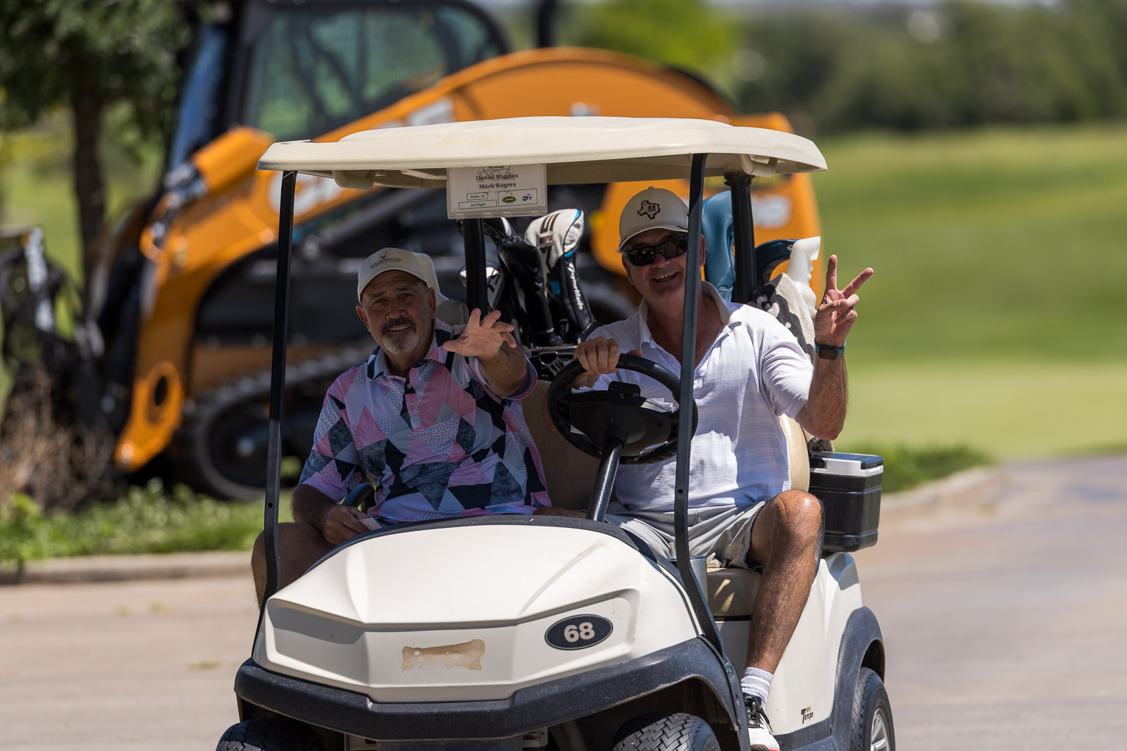 Two men riding in a white golf cart, waving at the camera on a sunny day with an orange construction vehicle in the background.