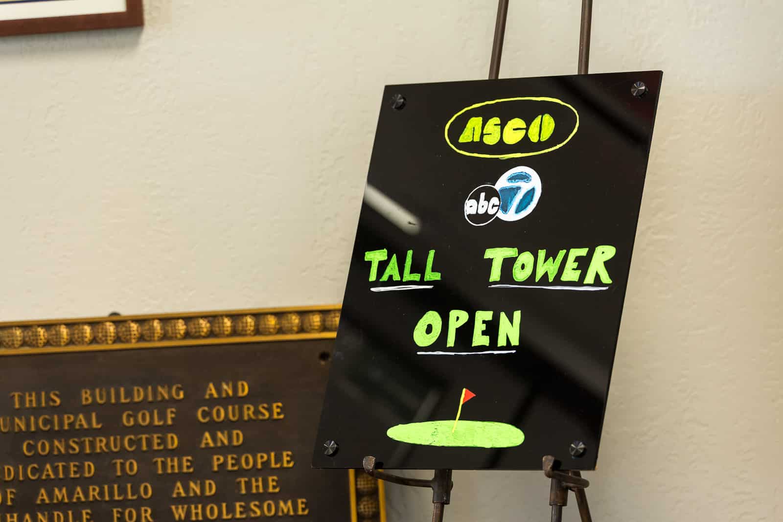 Black sign on an easel reading 'TALL TOWER OPEN' in neon green, with a yellow oval logo and abc7 logo above and a small green putting graphic at the bottom.