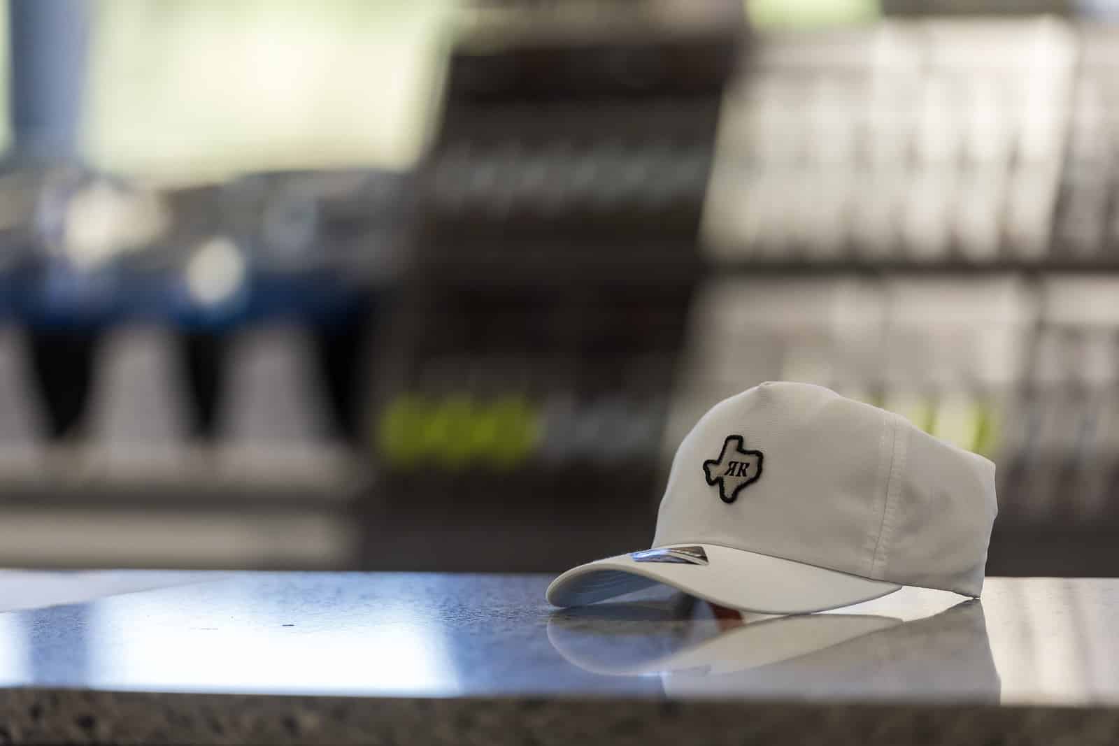 White baseball cap with a black Texas-shaped patch resting on a polished counter, blurred store shelves in the background.