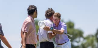 Two men on a sunny golf course hug as a third person looks on, holding golf clubs and wearing casual shirts.
