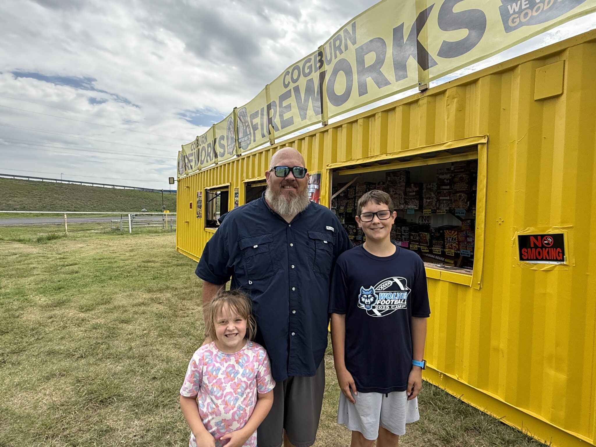 IMG_5647 | Press Pass Sports Man with sunglasses and a beard posing with two children outside a bright yellow fireworks stand on a cloudy day.