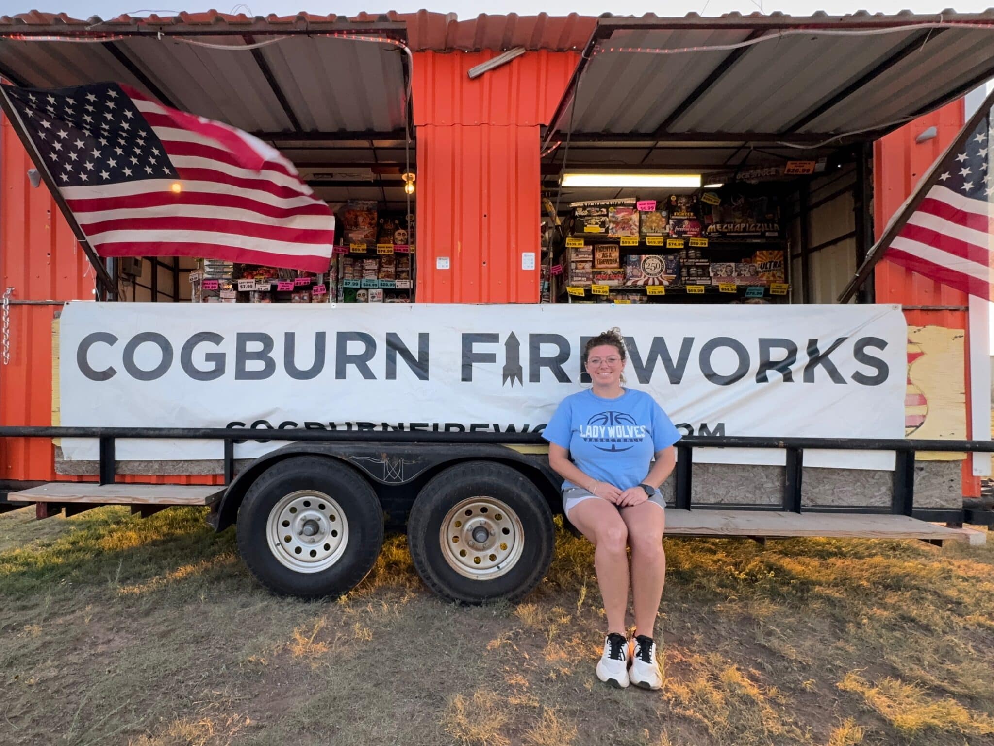 IMG_4432 | Press Pass Sports Woman in blue t-shirt sitting on a fireworks trailer in front of Cogburn Fireworks, flanked by American flags and a large banner.