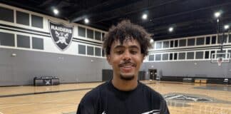 Smiling young man with curly hair in a black graphic T-shirt stands in a basketball gym with a Raiders emblem on the wall behind him.