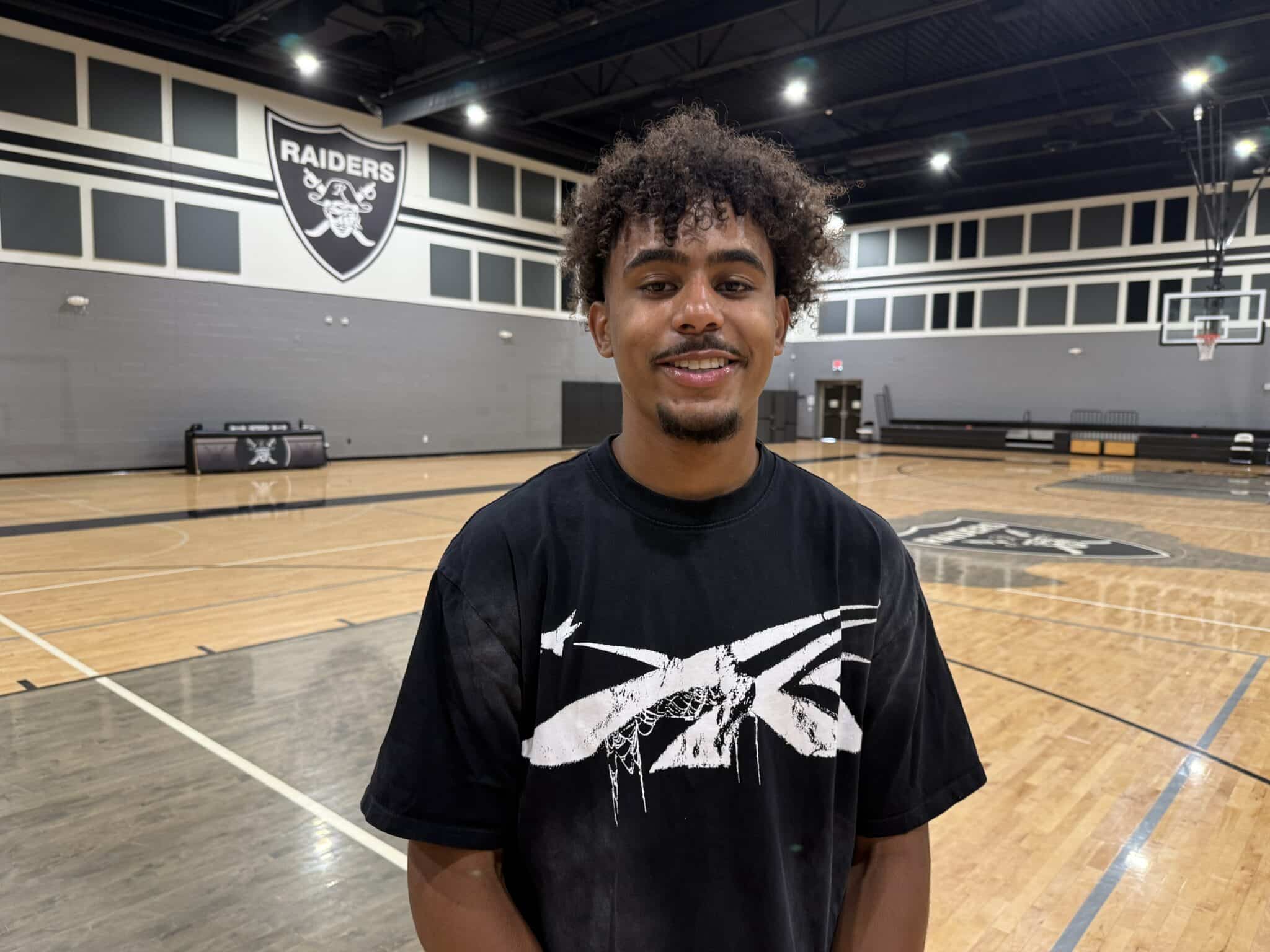 IMG_5641 | Press Pass Sports Smiling young man with curly hair in a black graphic T-shirt stands in a basketball gym with a Raiders emblem on the wall behind him.