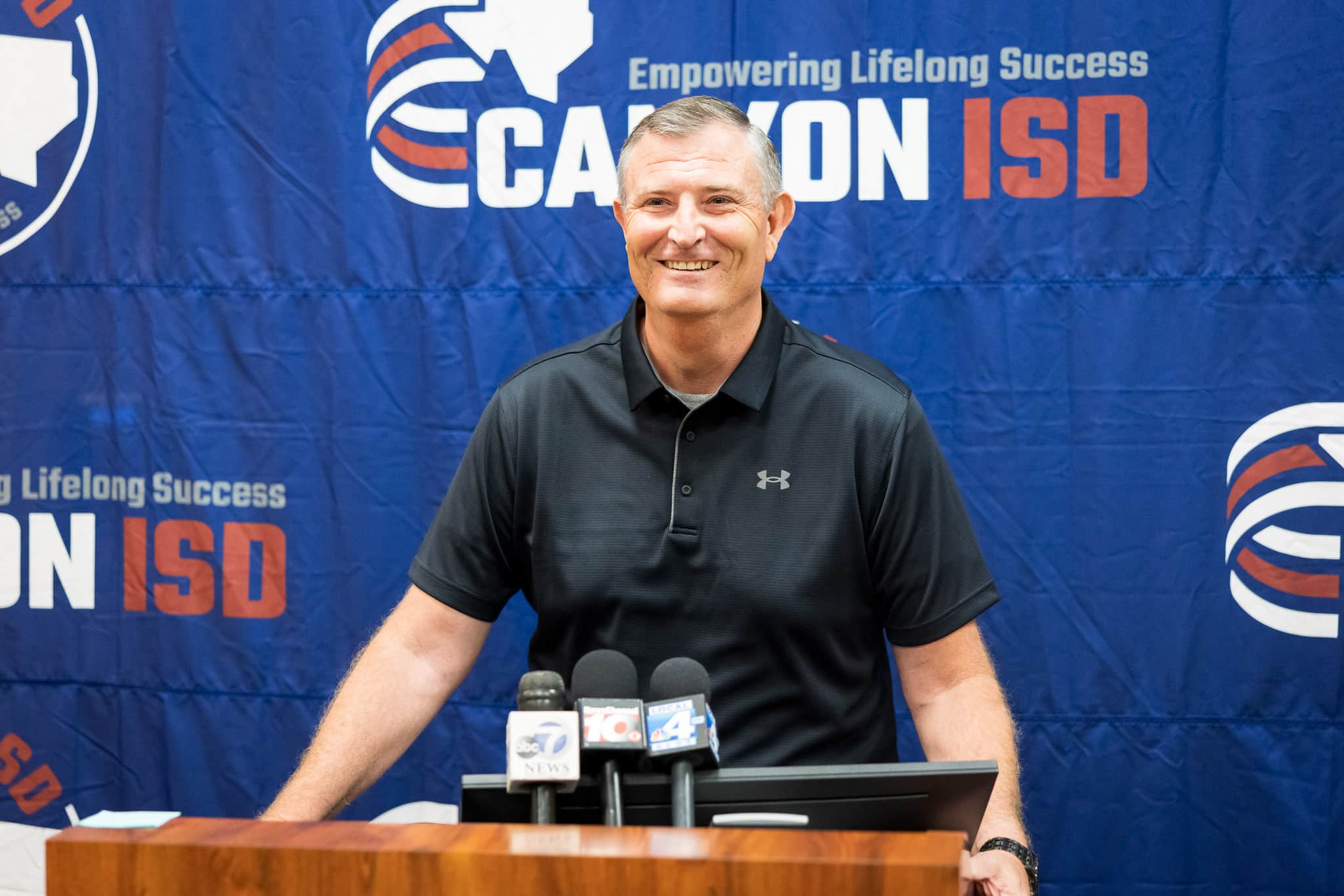 randall-basketball-coach-2 | Press Pass Sports Smiling man in a black polo speaks at a press conference, microphones on the podium, blue banner backdrop with logos behind him.