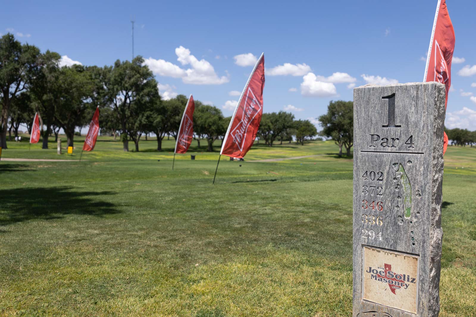 Stone marker for hole 1, Par 4, on a grassy golf course with red flags along the fairway and trees under a blue sky.
