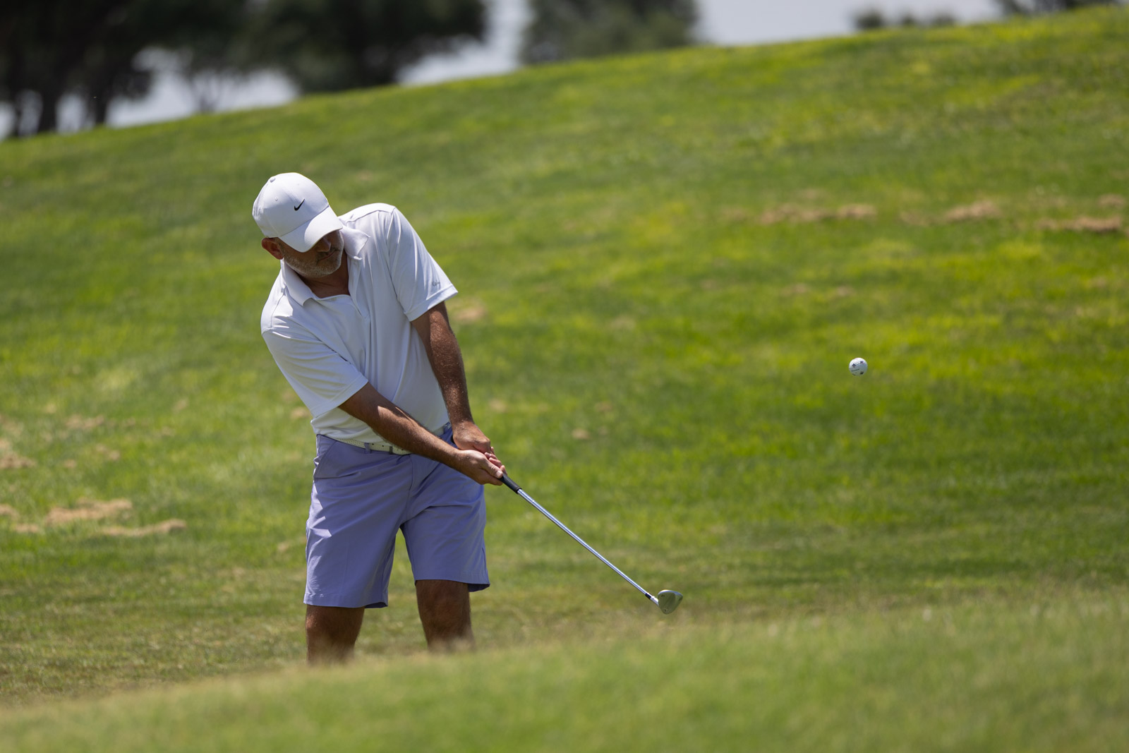 Man in a white polo and cap swinging a golf club on a sunny course, ball in mid-air to the right