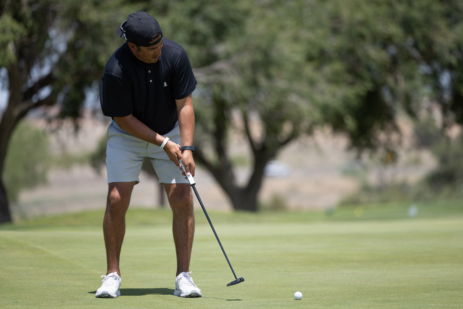 Golfer bending over to line up a putt on a green, wearing a black cap and polo with light shorts, holding a putter.