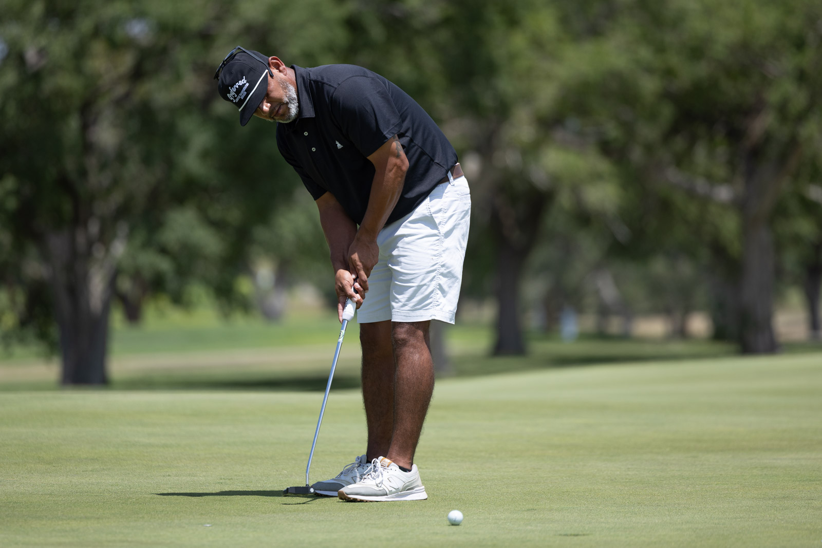 Golfer in a black polo and white shorts lining up a putt on a green fairway under sunny skies