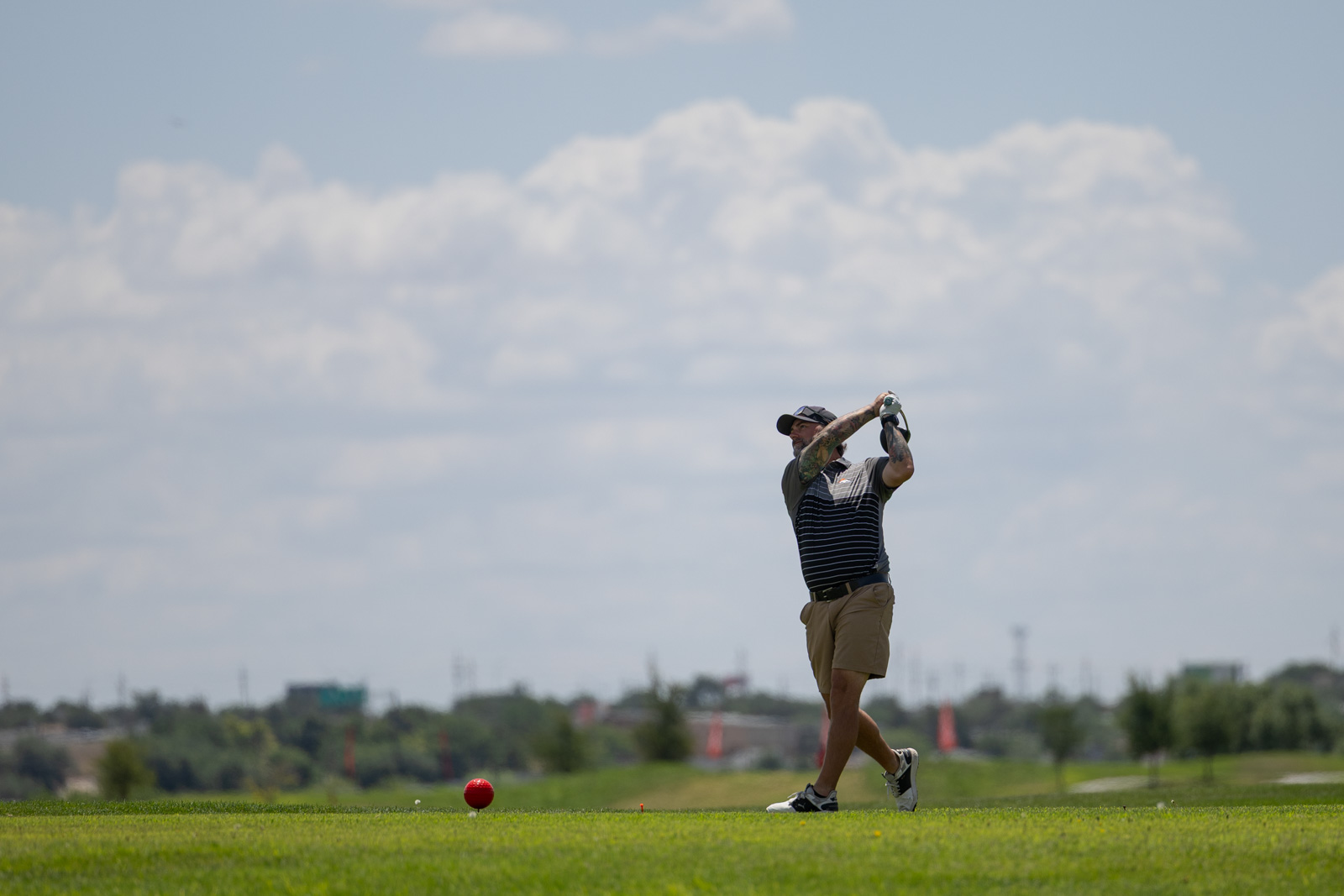 Man in a striped polo and beige shorts mid-swing on a sunlit golf course, with a red ball on the tee.