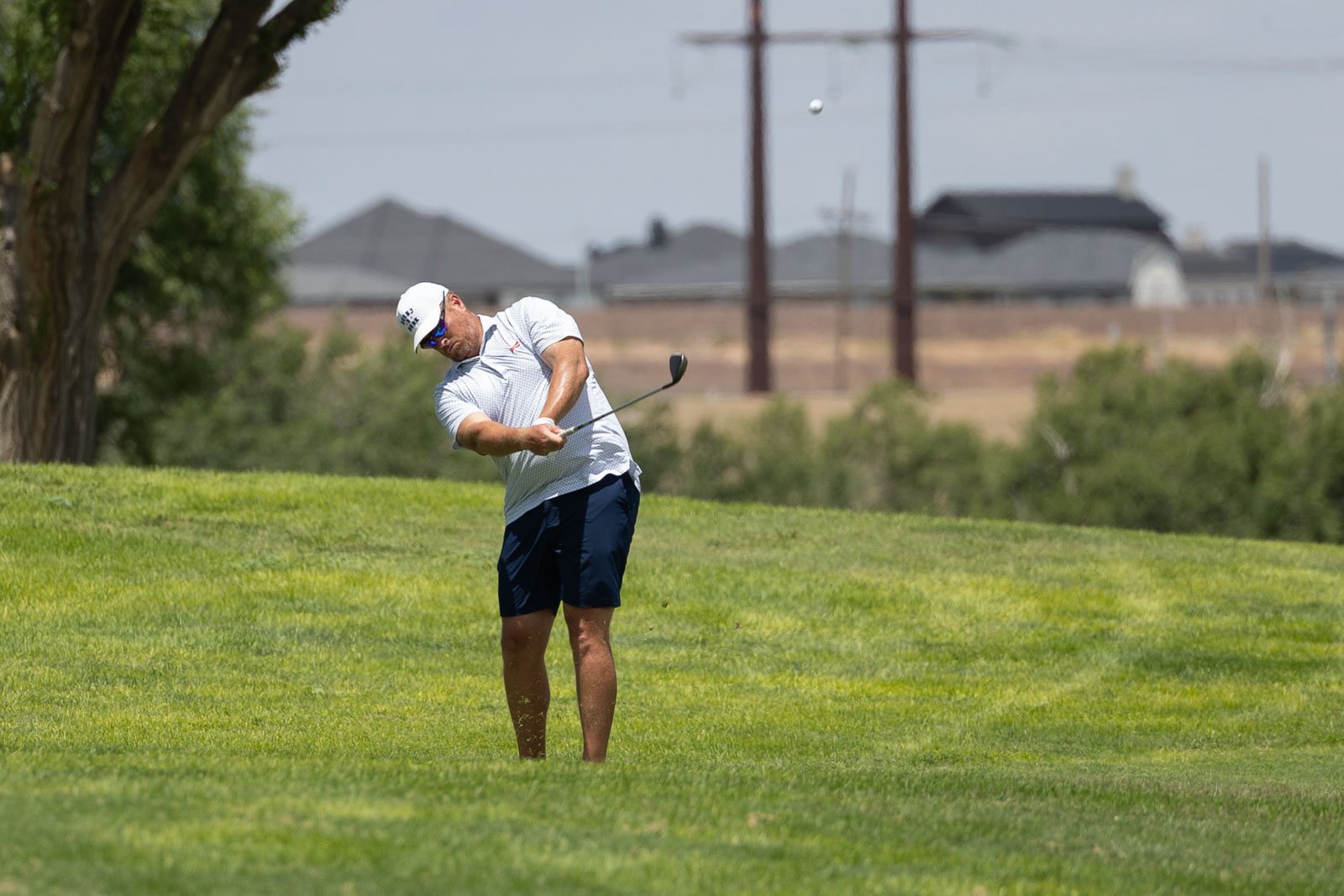 A male golfer in a white polo and navy shorts swings a club on a sunny green fairway, ball in midair behind him.