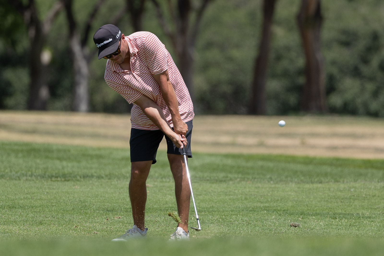 Male golfer in a striped polo and cap preparing a golf swing on a well-manicured fairway, ball in midair nearby