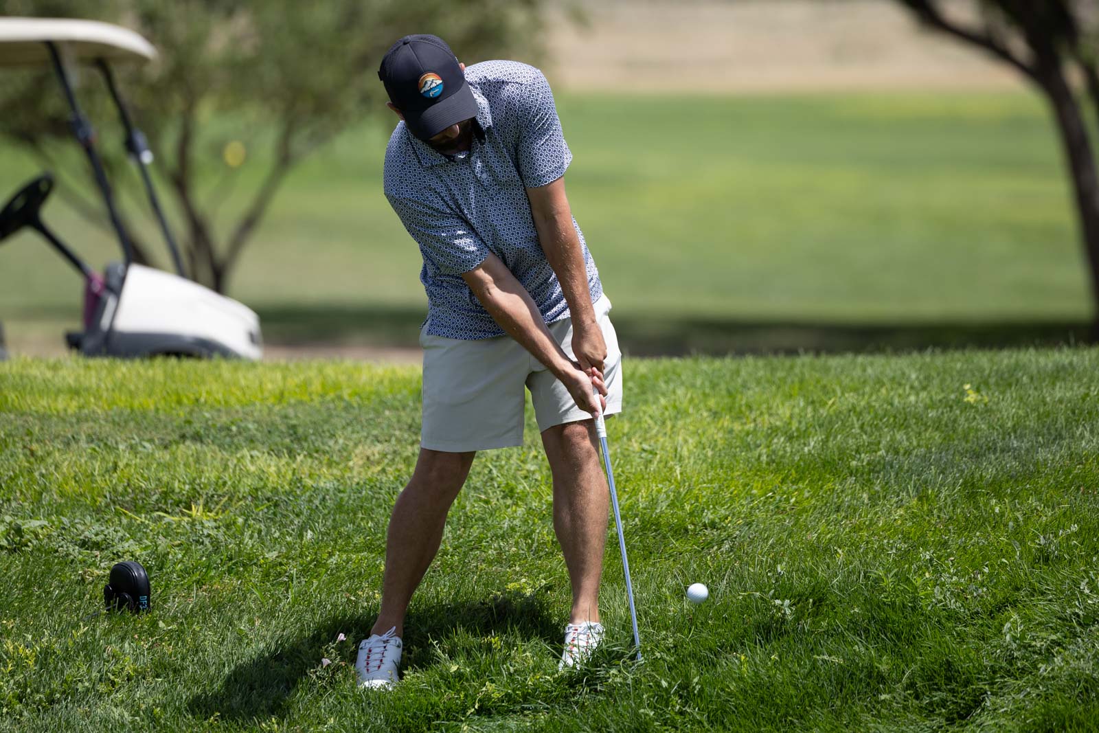Man in a patterned polo and light shorts swinging a golf club on a sunny golf course, ball teed up on the grass with a golf cart in the background.