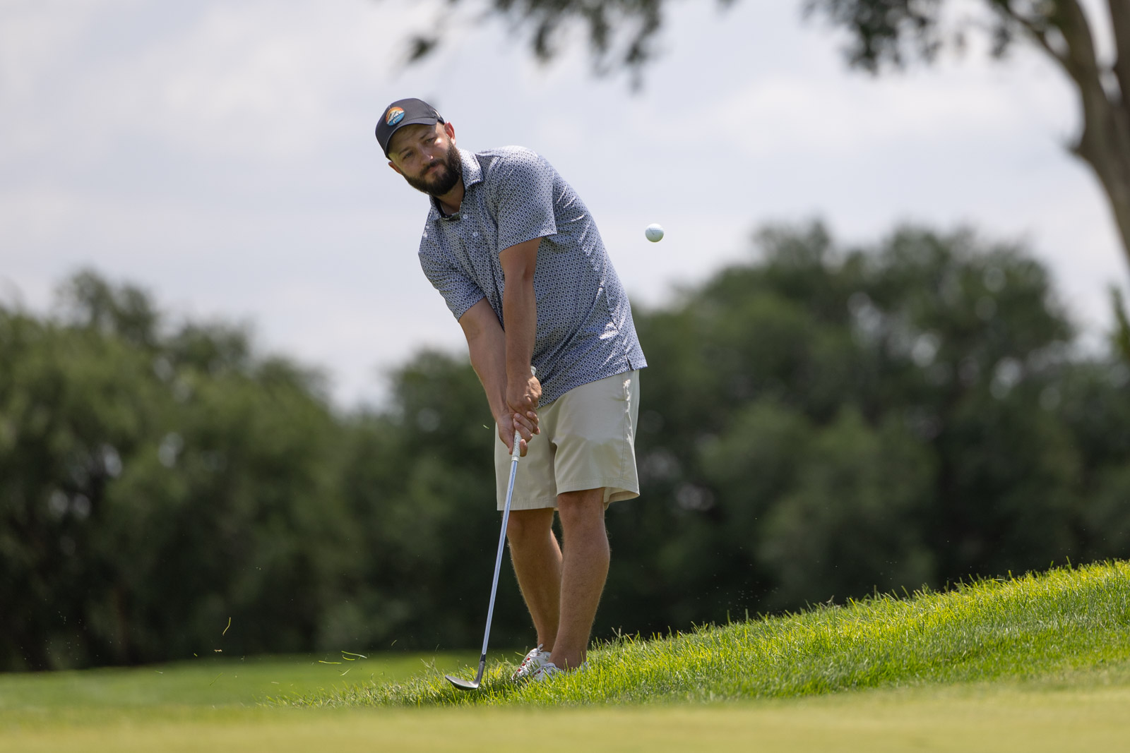 Man in a cap and patterned shirt swings a golf club on a sunny course, a golf ball midair nearby on a grassy slope