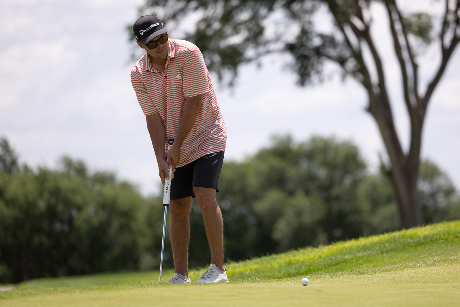 Golfer in a red-striped polo prepares to putt on a green putting surface with a white ball nearby.