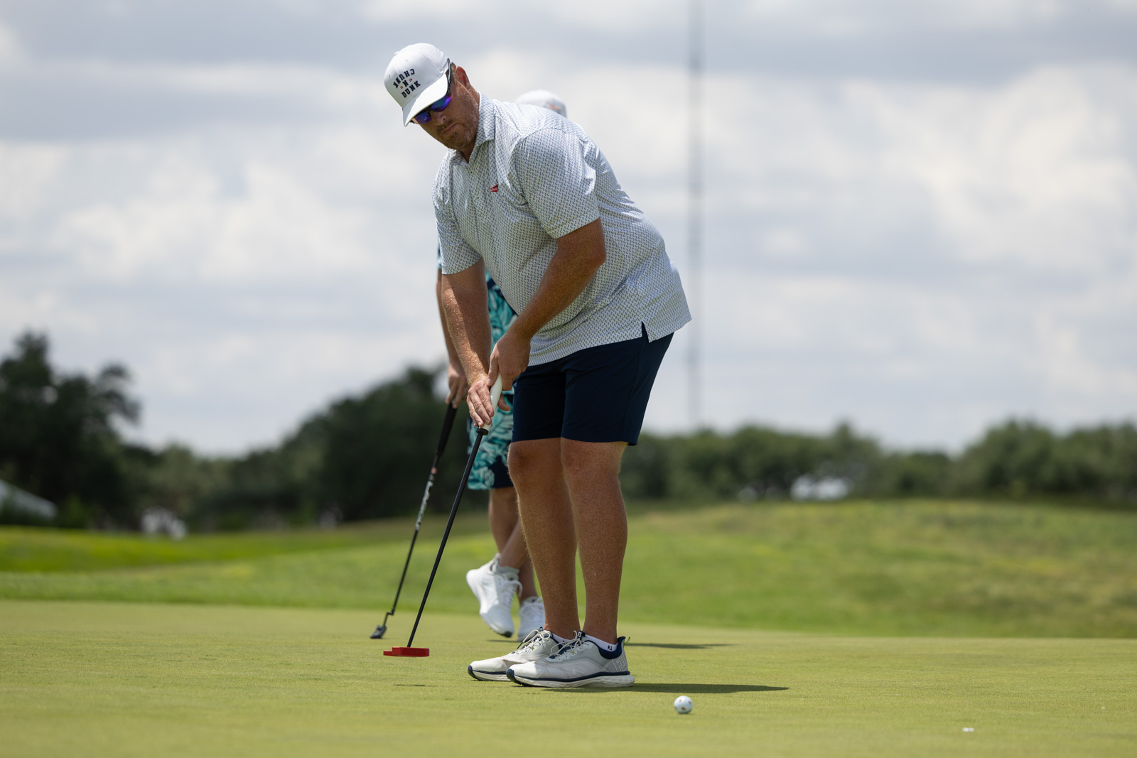 A golfer bending over to putt on a green, with a putter and golf ball nearby under a sunny sky