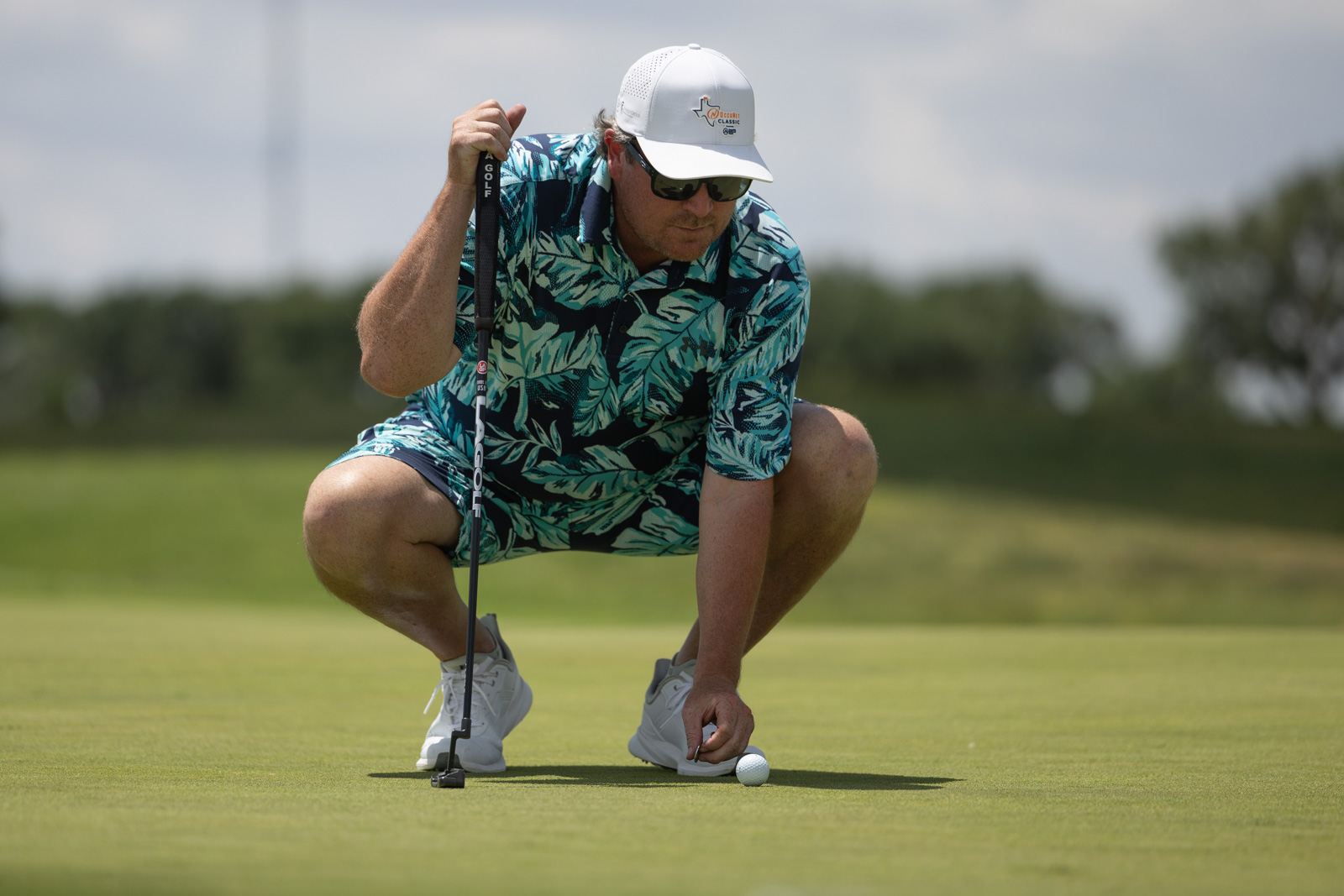 Golfer crouches on the green, lining up a putt with a black putter, wearing a tropical print shirt and white cap.