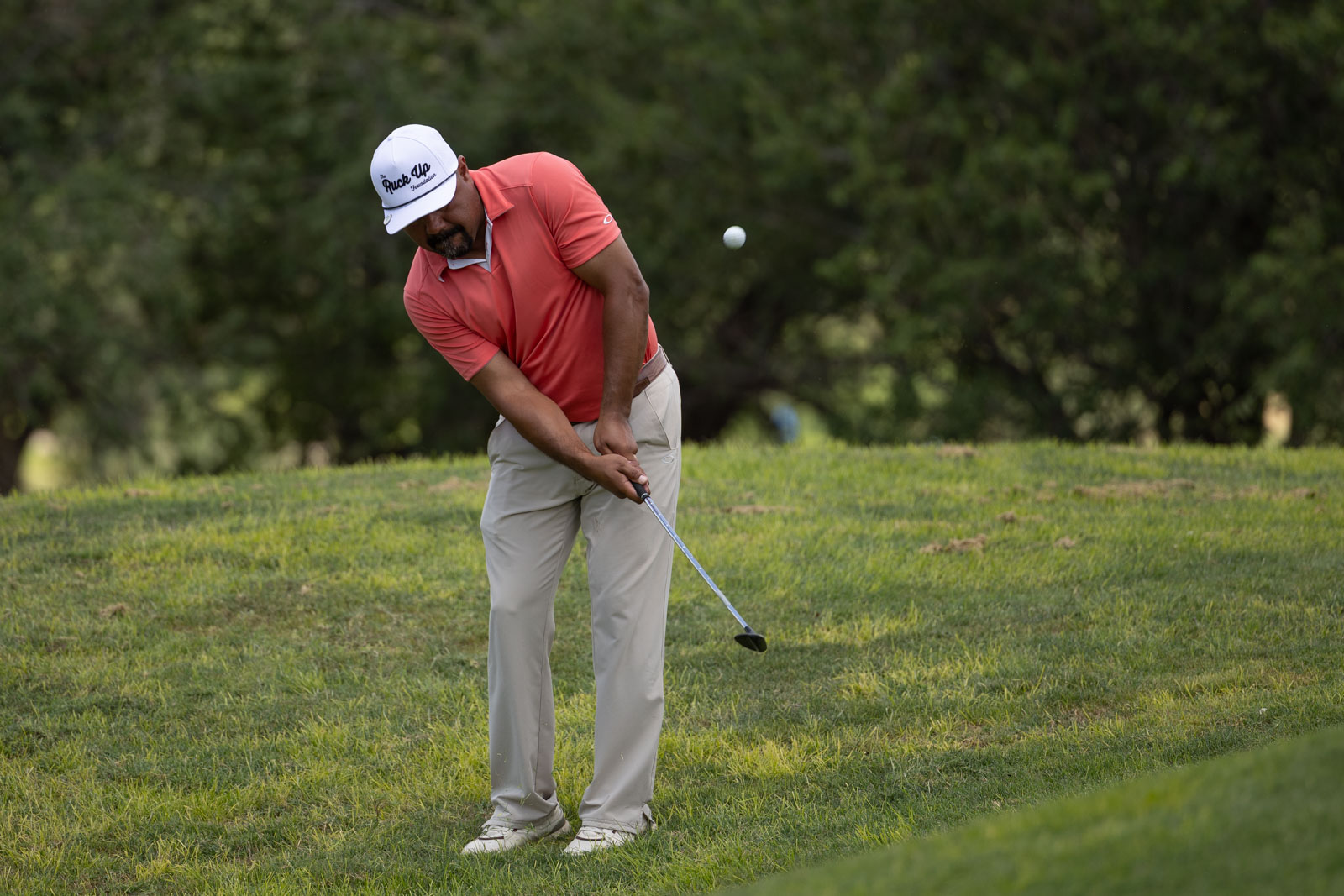 Golfer in a coral polo and white cap swinging a club on a grassy course as a ball flies through the air between trees in the background.
